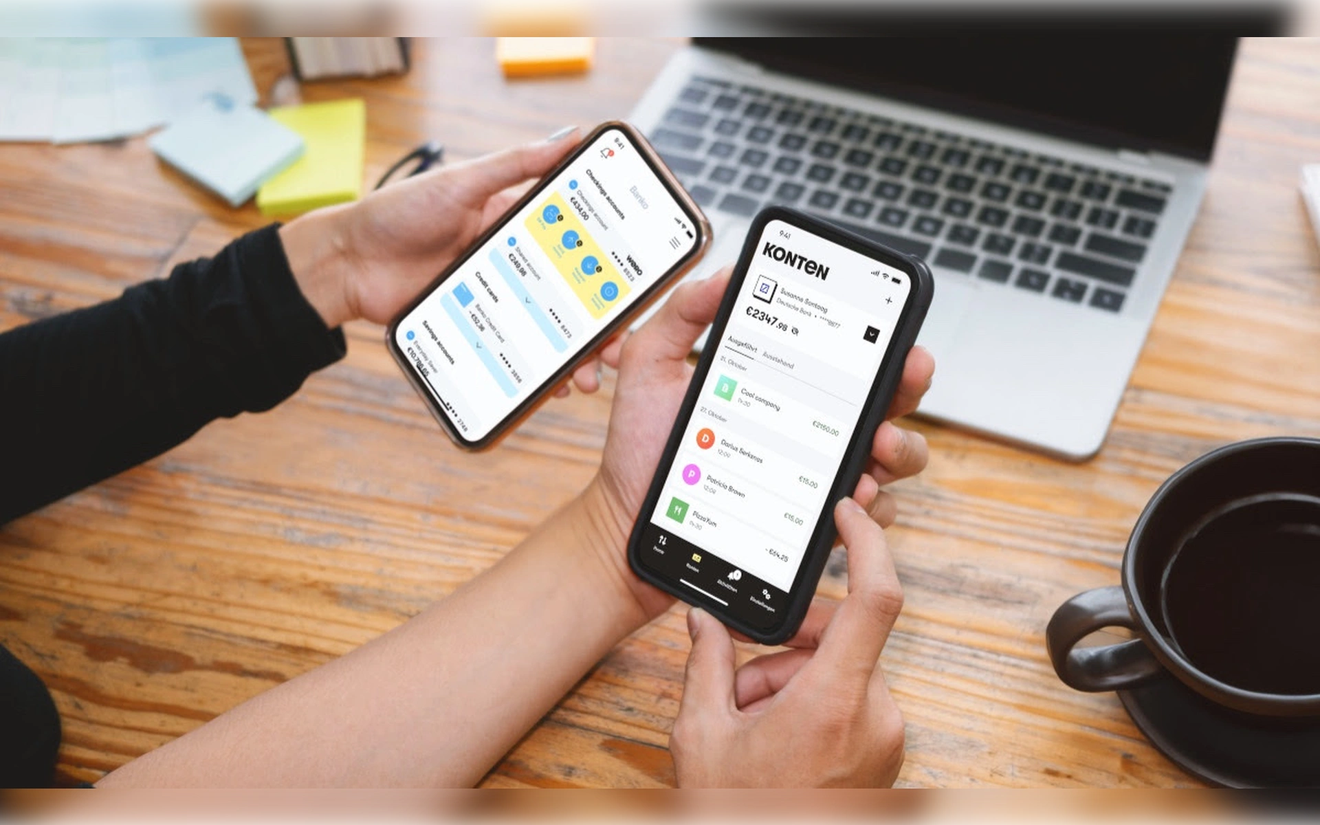 Zwei Personen halten Smartphones über einem Holztisch und nutzen Banking-Apps (Wero/Konten) für Überweisungen und Finanzübersichten. Im Hintergrund ein Laptop.