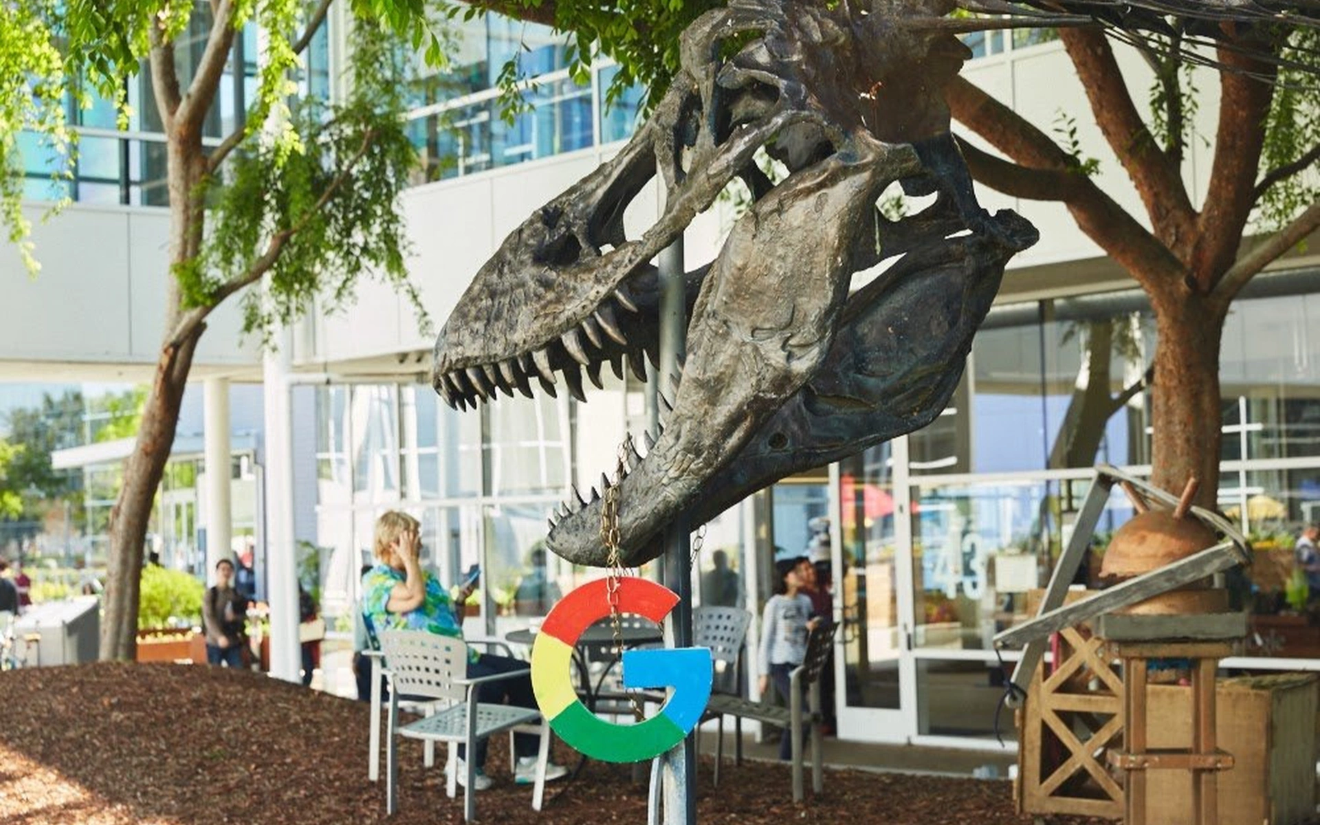 Ein großer Dinosaurier-Schädel (T-Rex) aus Metall mit einem hängenden, bunten Google-Logo vor einem Bürogebäude auf dem Googleplex-Campus.