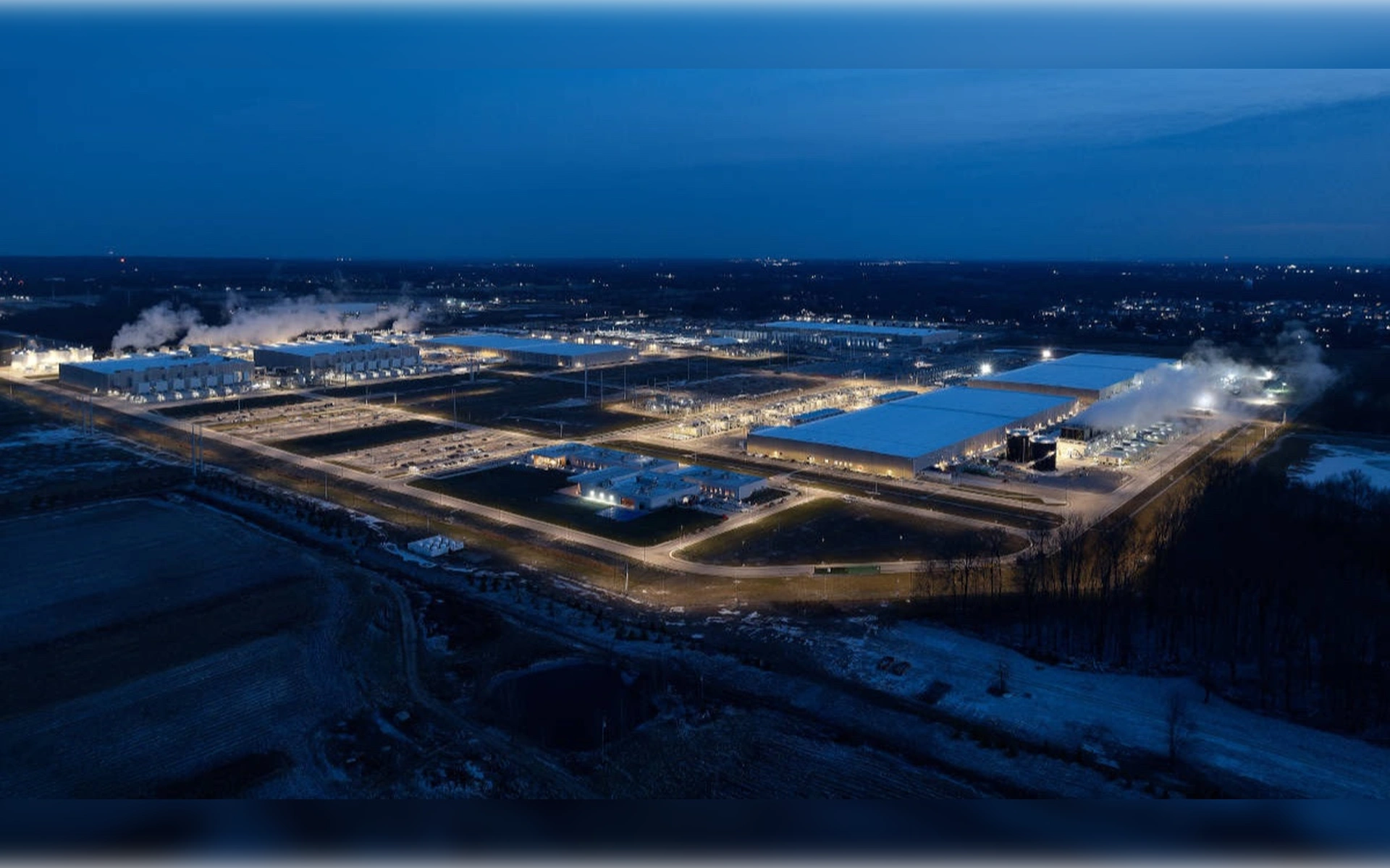 Eine nächtliche Luftaufnahme eines riesigen, hell erleuchteten Google-Rechenzentrumskomplexes in New Albany mit rauchenden Kühltürmen inmitten einer dunklen Landschaft.