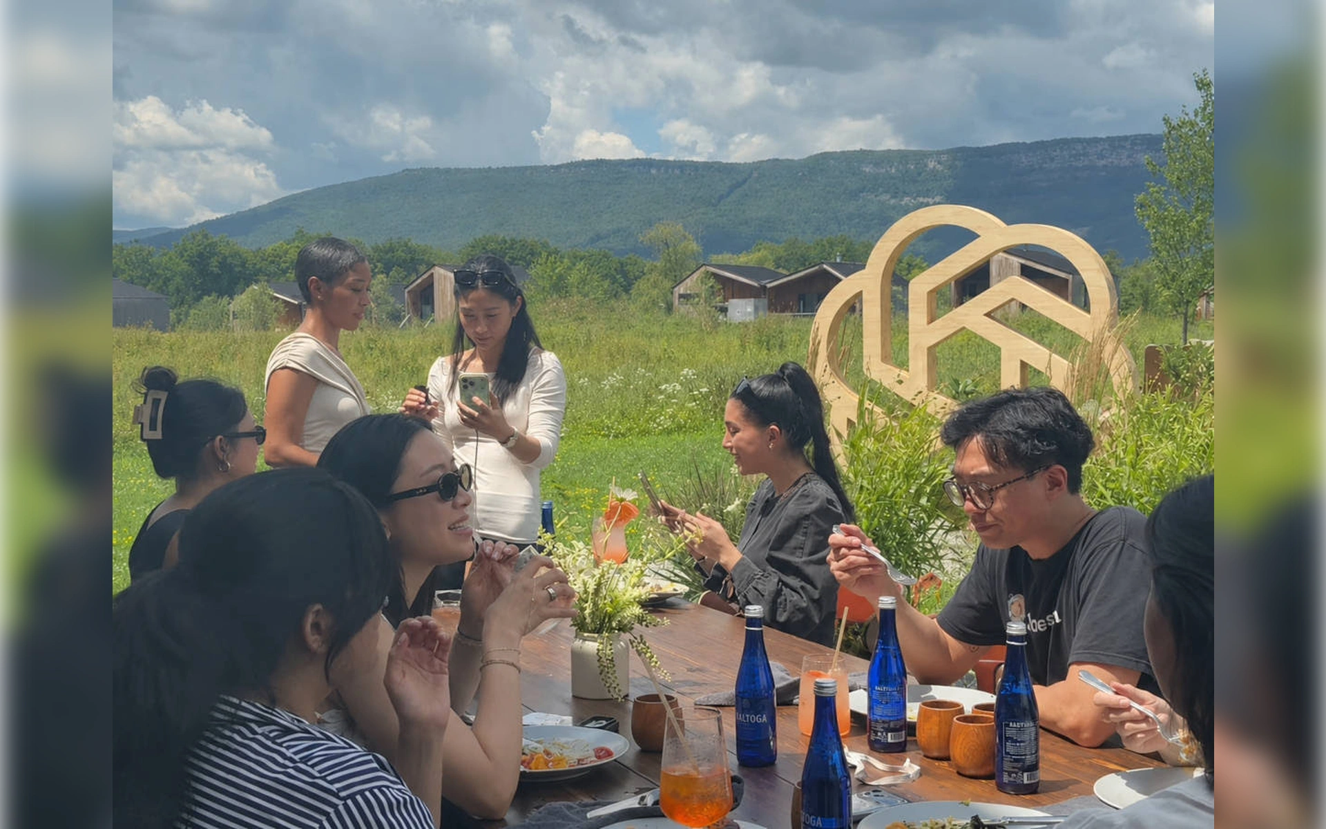 Eine Gruppe von Menschen sitzt im Freien an einer langen Festtafel beim Essen. Im Hintergrund ist eine große Holzskulptur des OpenAI-Logos vor Bergen zu sehen.