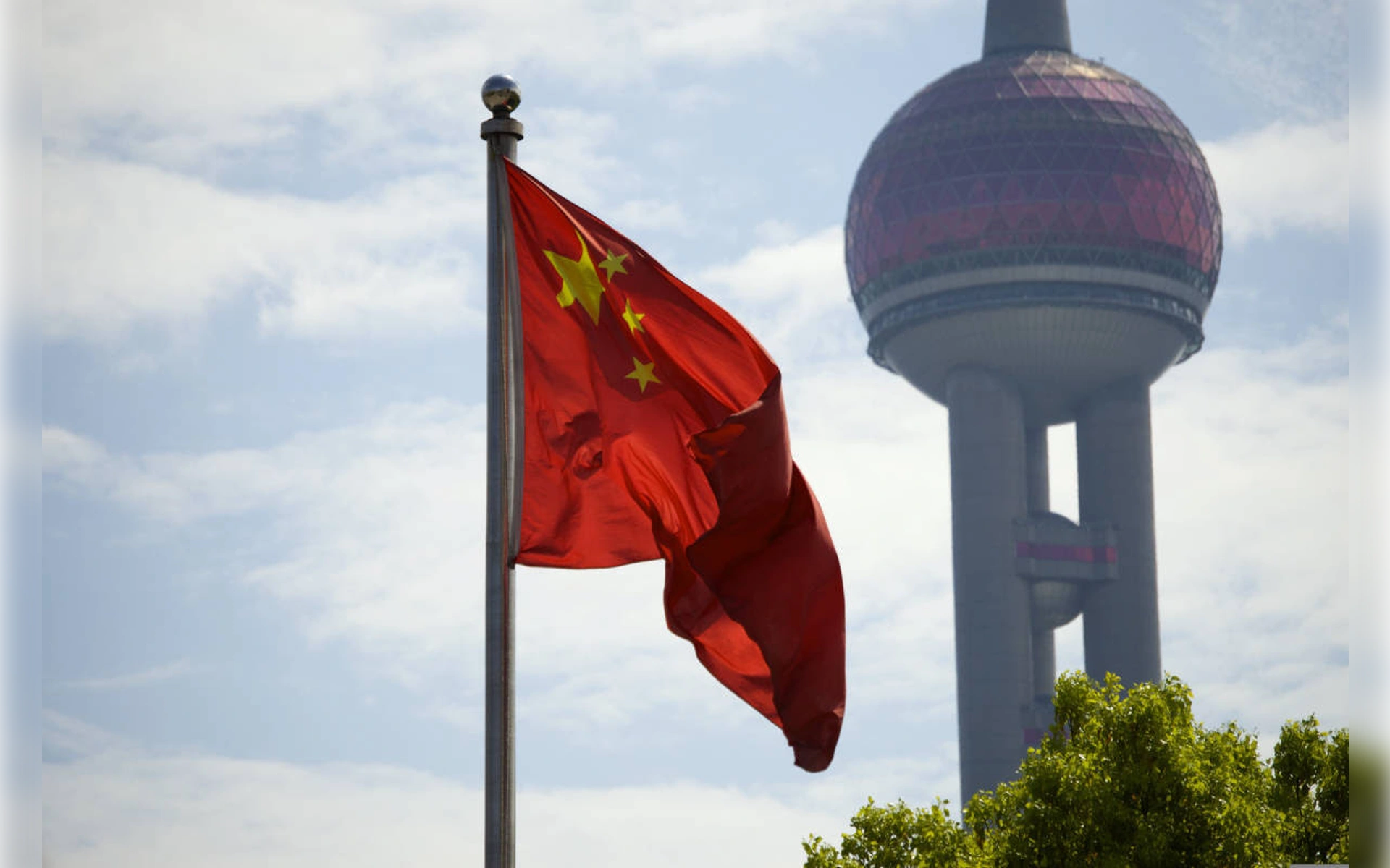 Die rote chinesische Nationalflagge weht an einem Masten vor dem leicht unscharfen Hintergrund des Oriental Pearl Tower in Shanghai unter blauem Himmel.