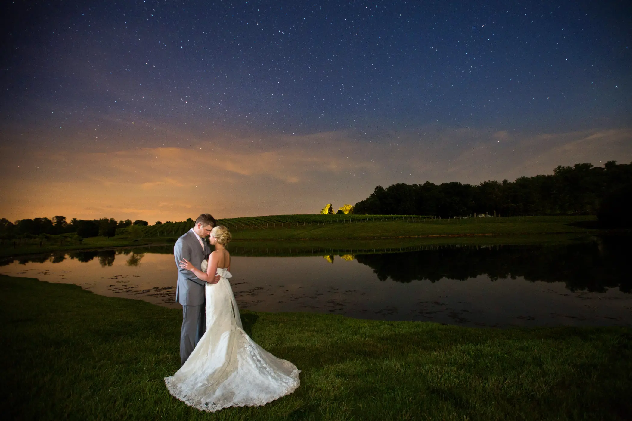 A bride and groom embrace outdoors near a calm body of water at dusk, with a darkening sky and trees in the background.