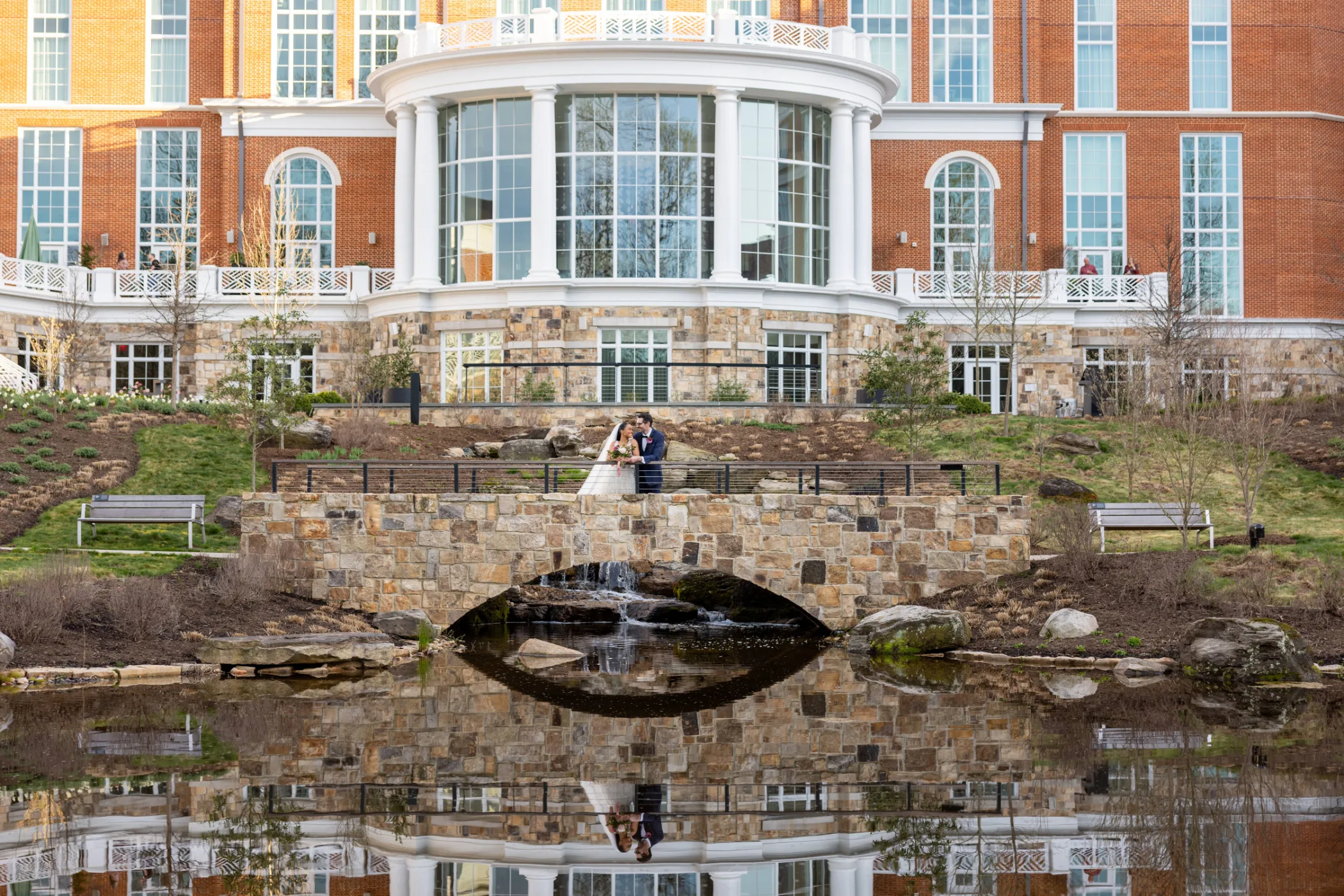 A bride and groom embrace and smile outdoors near an orange Aperol Spritz cart and a bouquet of flowers.