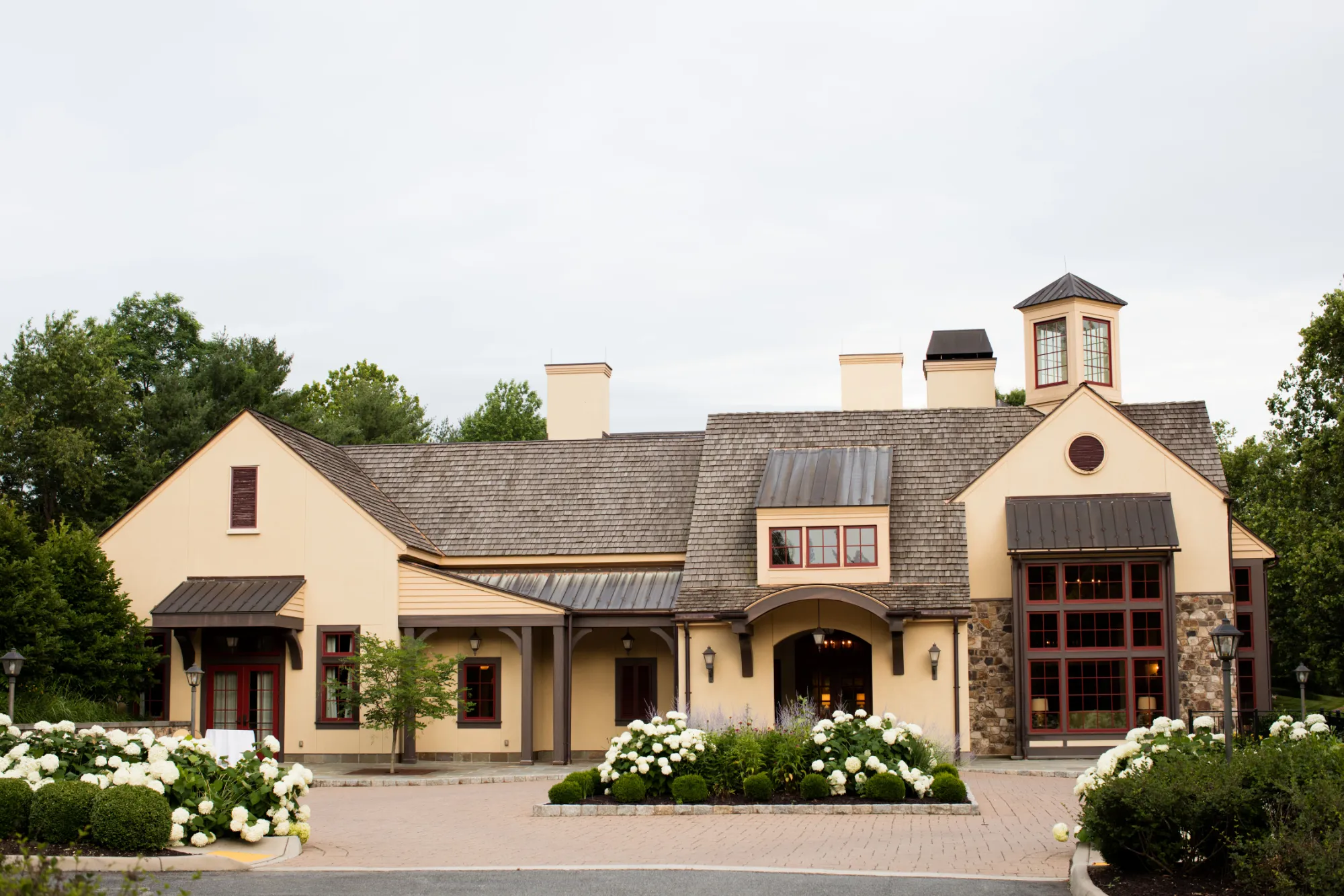 Large beige house with multiple chimneys, gabled roofs, and a landscaped front yard, surrounded by trees under a cloudy sky.
