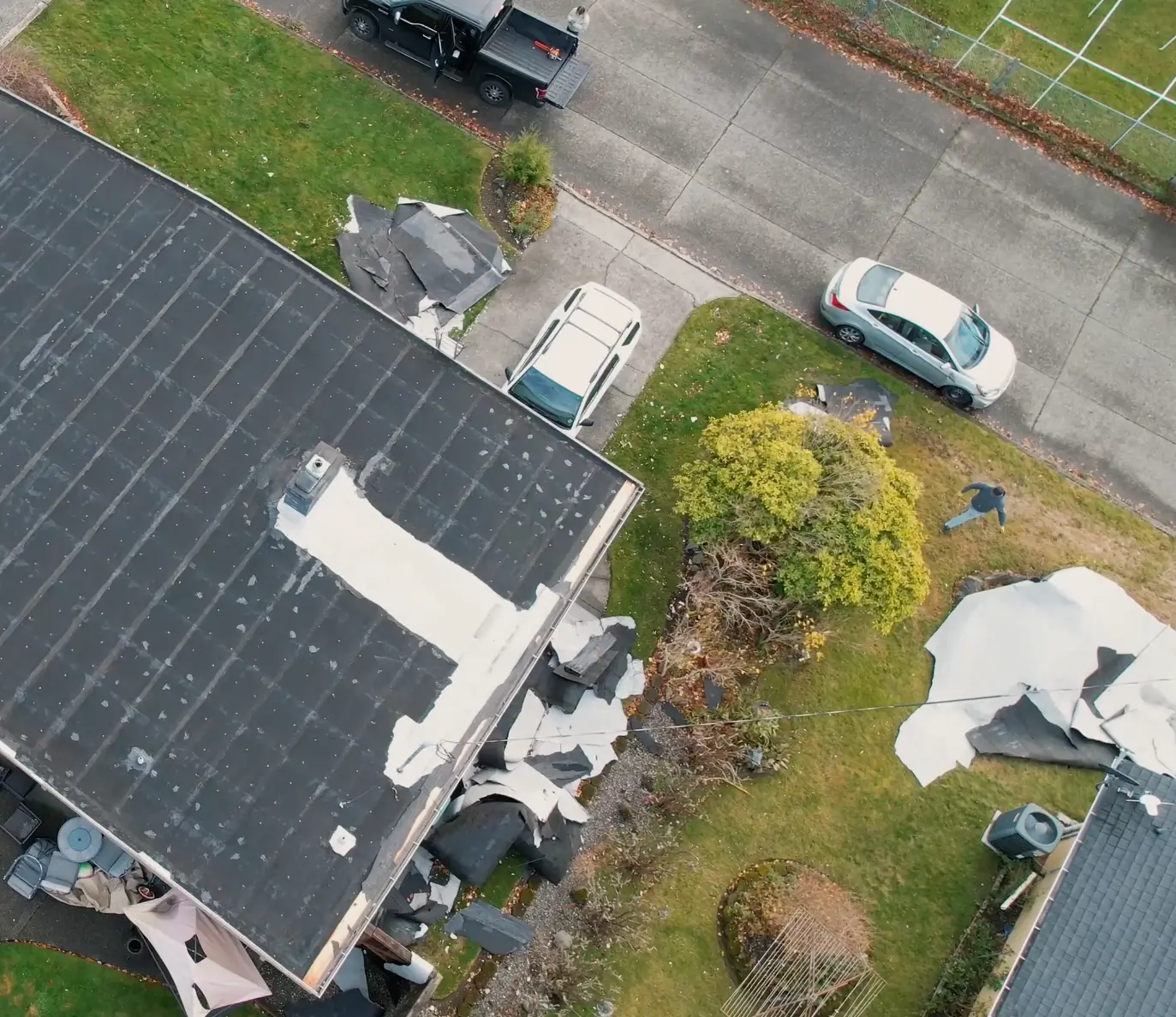 "Flat roof with widespread damage, torn roofing membrane scattered across the yard, and patchwork repairs indicating wind damage and material failure.
