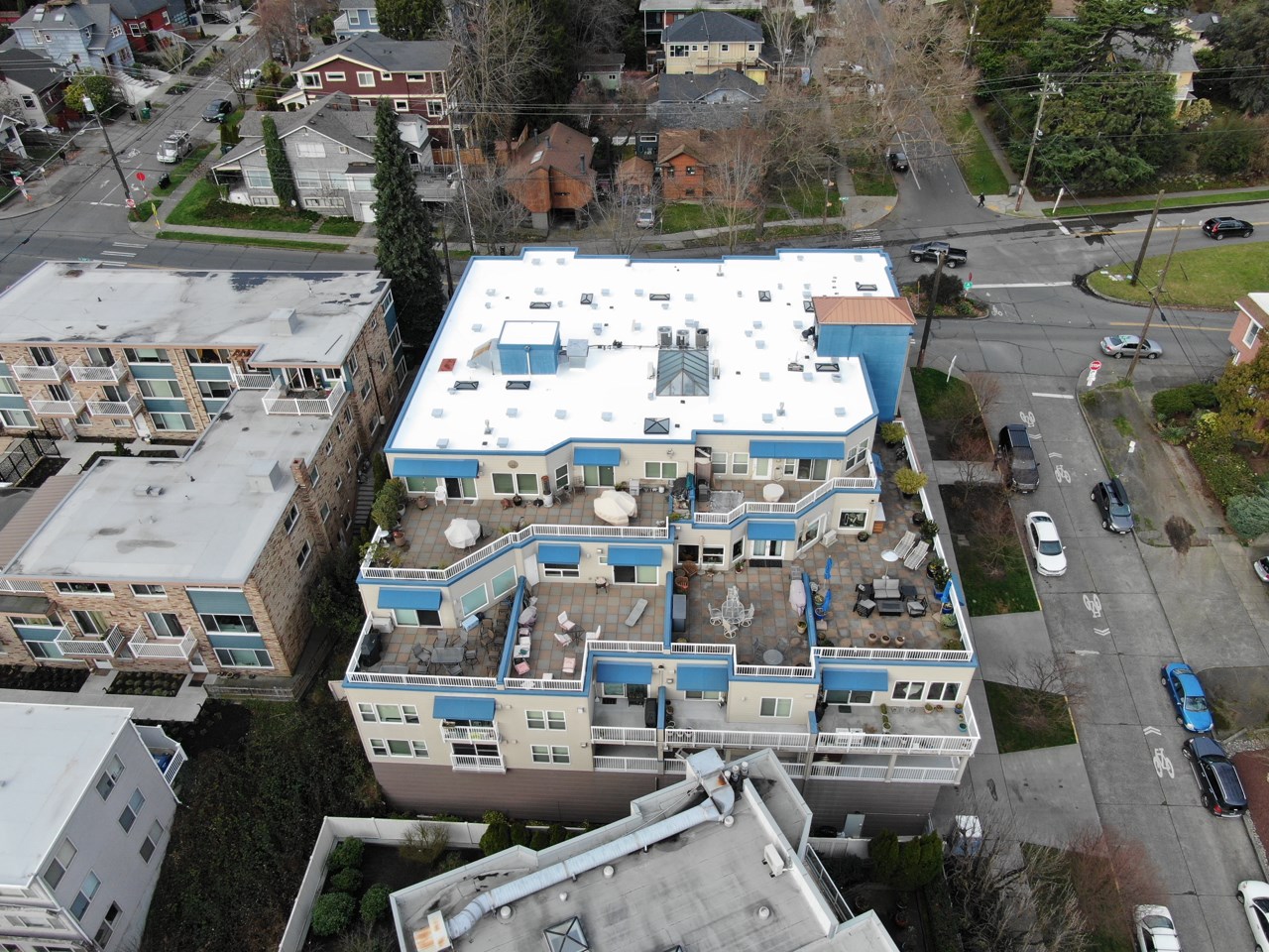 Bird's-eye view of a completed white silicone roof restoration on a multi-family residential building, surrounded by older flat roofs in a dense urban neighborhood.