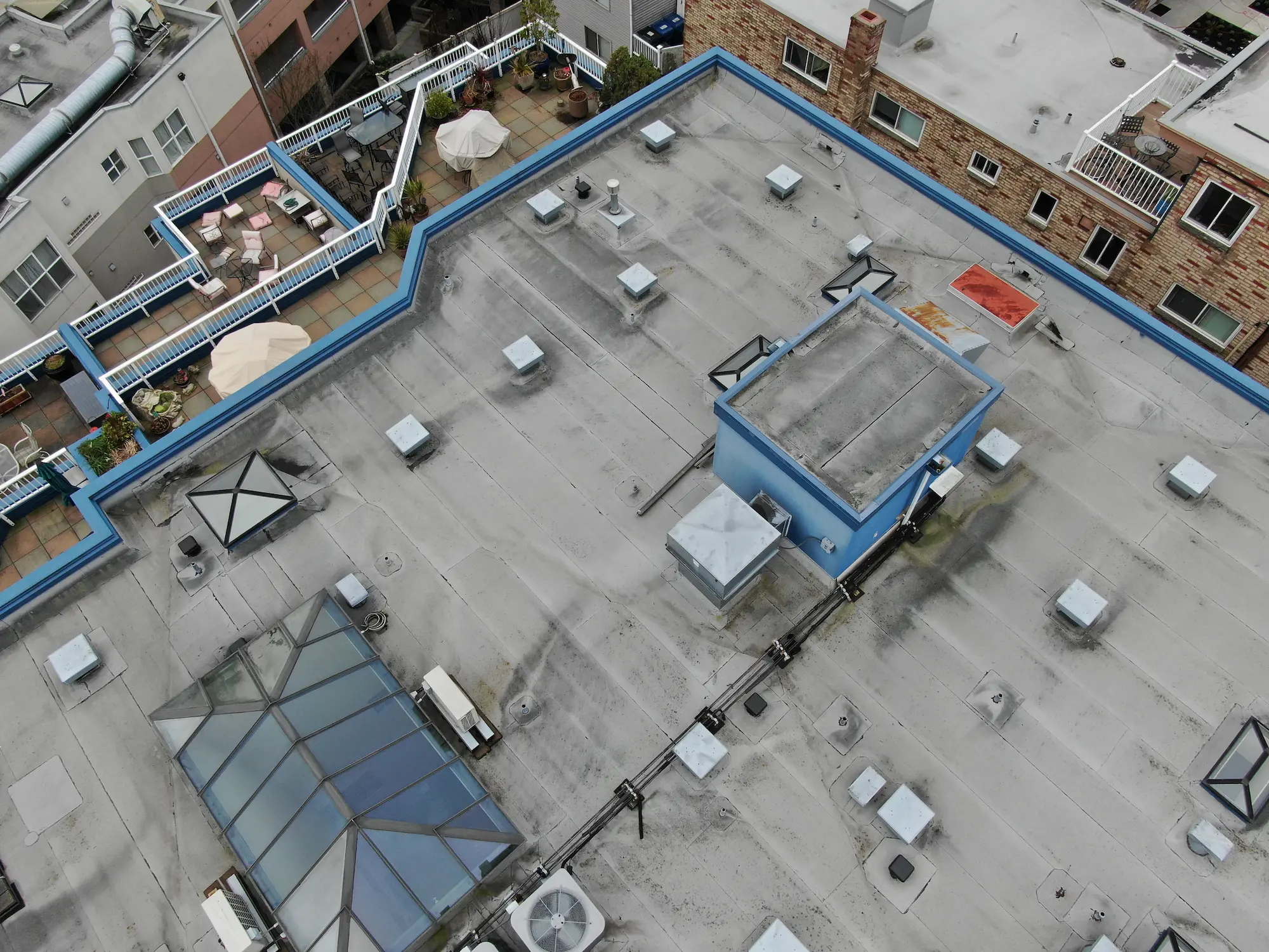 Aerial close-up of an aging flat roof on a multi-family building, showing membrane wear, skylights, HVAC units, and rooftop patios with umbrellas and seating areas.