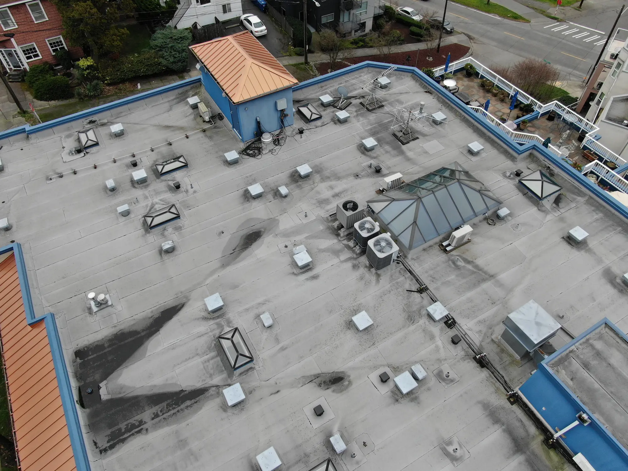 Aerial view of an aged commercial flat roof with worn membrane, visible water staining, skylights, and rooftop HVAC units on a multi-family building.