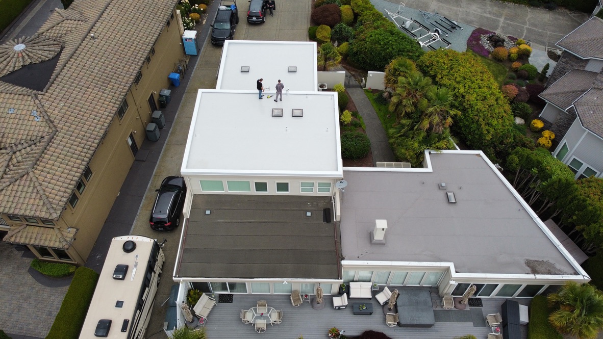 Drone view of residential home with newly installed white PVC flat roof next to older dark membrane roofing, highlighting the contrast between restored and aging roof sections.