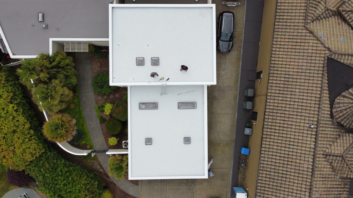 Aerial view of residential home with newly installed white PVC flat roofing system, featuring skylights, clean seams, and improved waterproofing in a landscaped neighborhood.