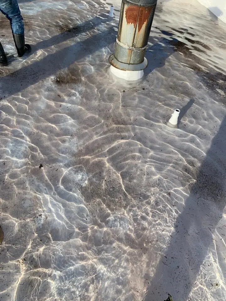 Close-up of flat roof with significant ponding water around a rusty vent pipe, indicating drainage issues and potential membrane damage requiring repair or restoration.
