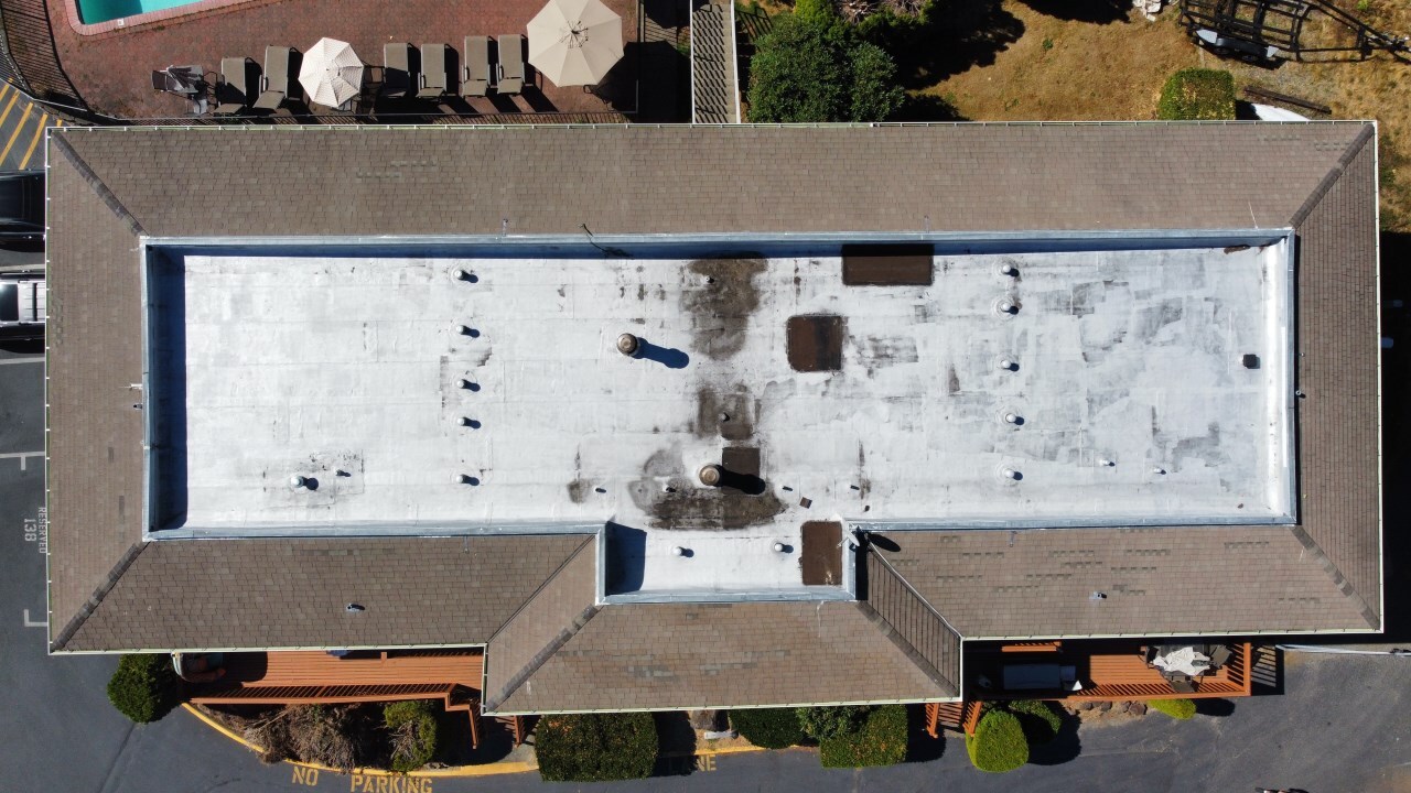 Top-down view of flat roof on a multi-unit building with visible staining, vent penetrations, and surrounding shingle roofing—indicating wear and potential need for restoration or coating.