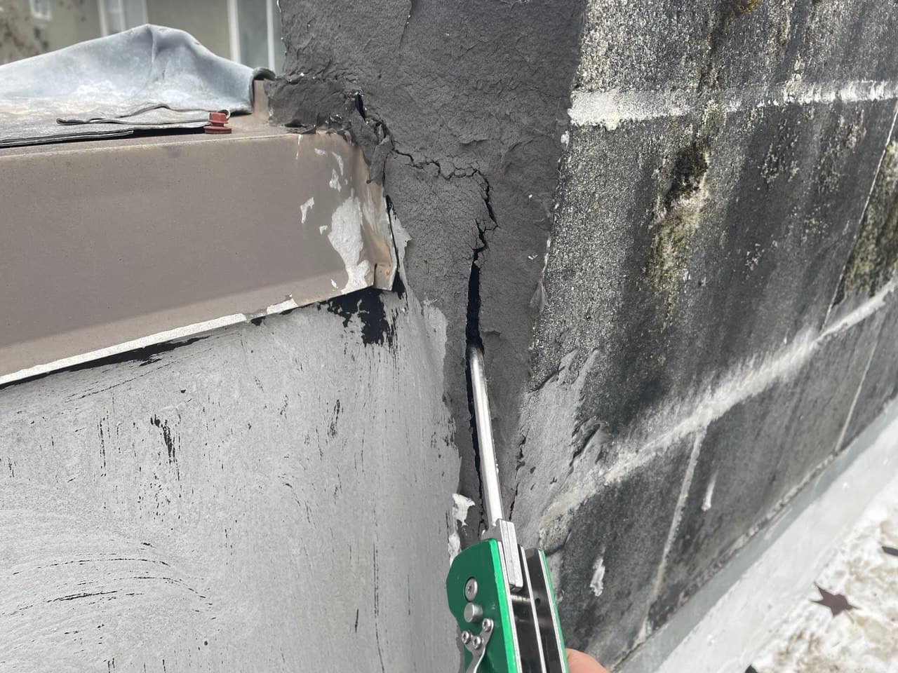 Cracked and deteriorating concrete wall around rooftop parapet, showing deep gaps and exposed flashing near metal cap flashing. Close-up of structural damage with moisture stains and signs of water intrusion, emphasizing the need for professional roof repair and waterproofing solutions.