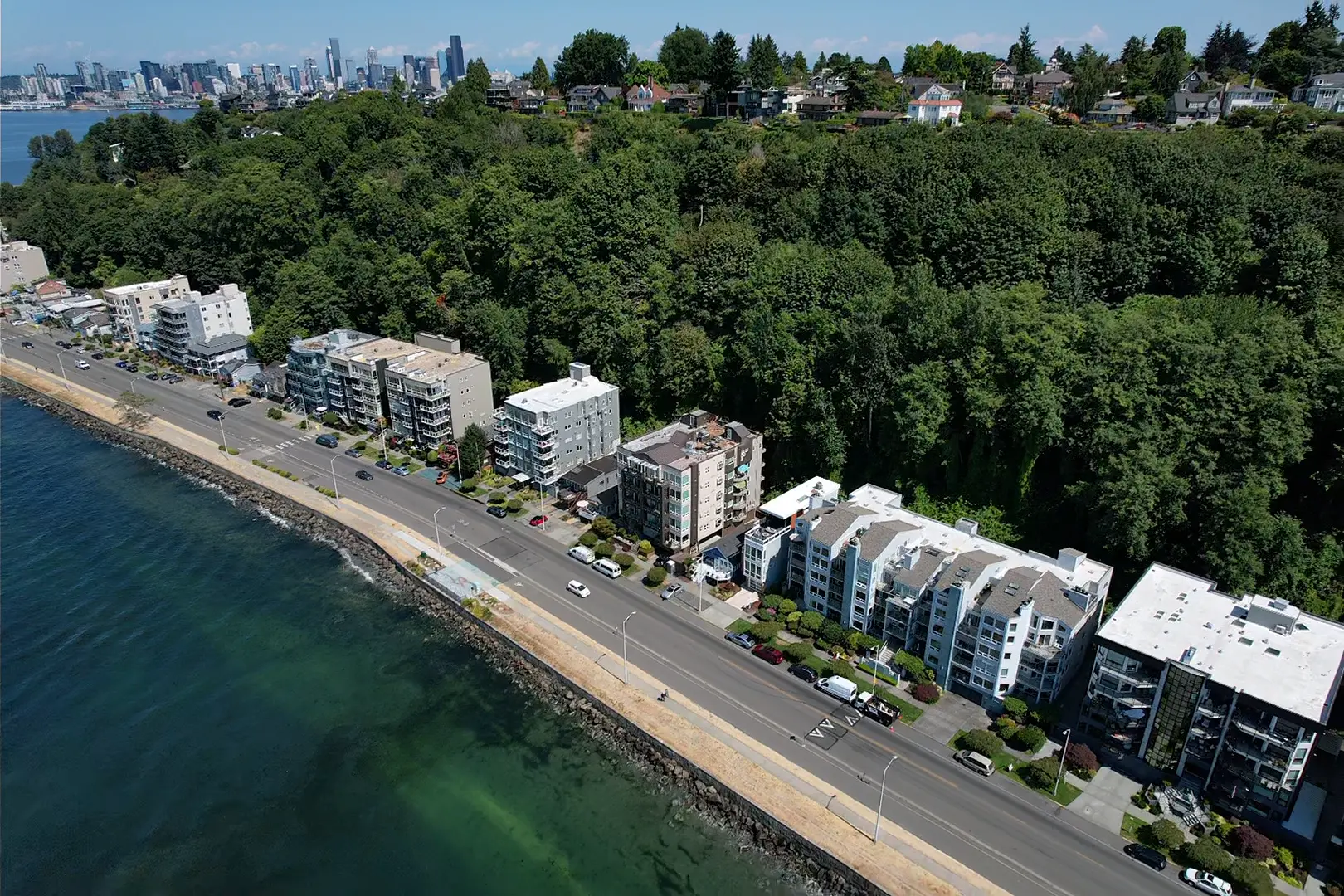 Waterfront apartments in West Seattle overlooking Elliott Bay, with downtown Seattle skyline views and lush green hillside surroundings, prime coastal real estate near Alki Beach.
