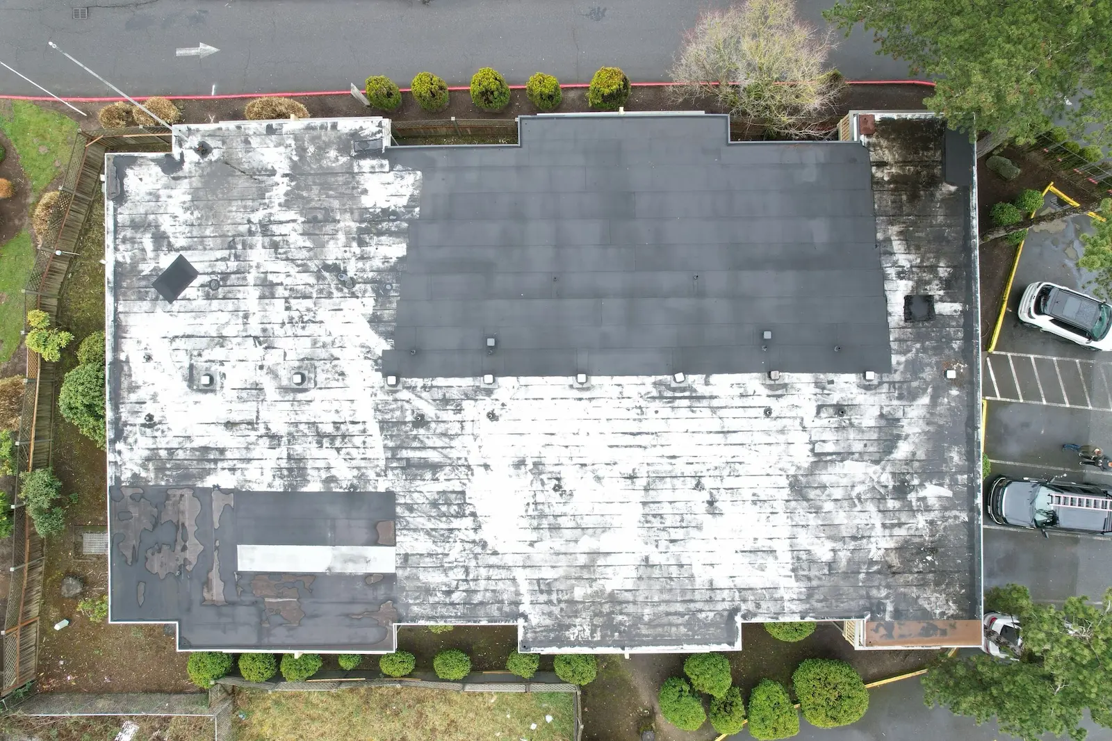 Aerial view of a multifamily building roof showing old patchwork repairs across a black roof section surrounded by a heavily weathered white membrane.