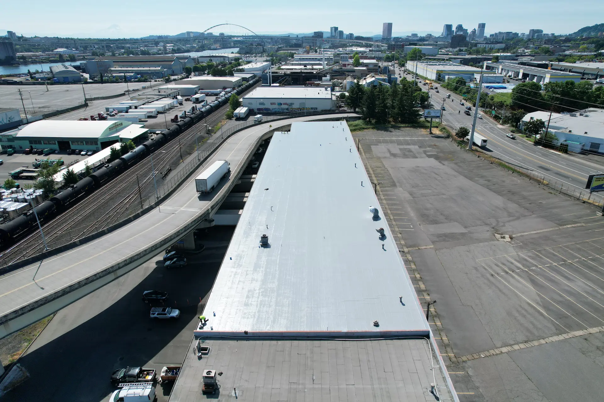 Drone view of a fully restored commercial flat roof with a newly applied gray silicone coating system on a long industrial warehouse beside a highway overpass.