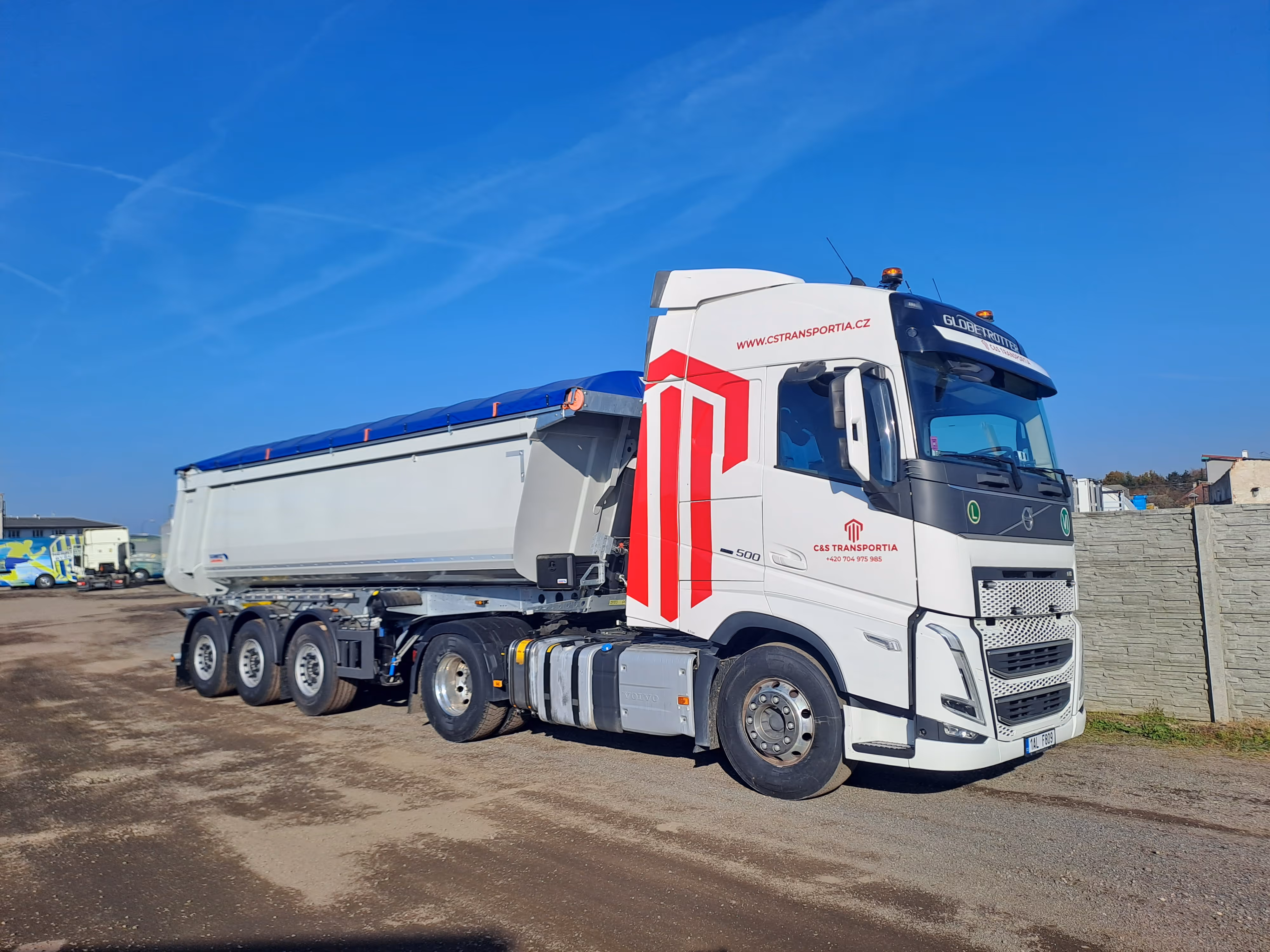 Bílý nákladní automobil s modrým plachtou a logem C&S Transportia zaparkovaný na šedém povrchu pod jasně modrou oblohou.