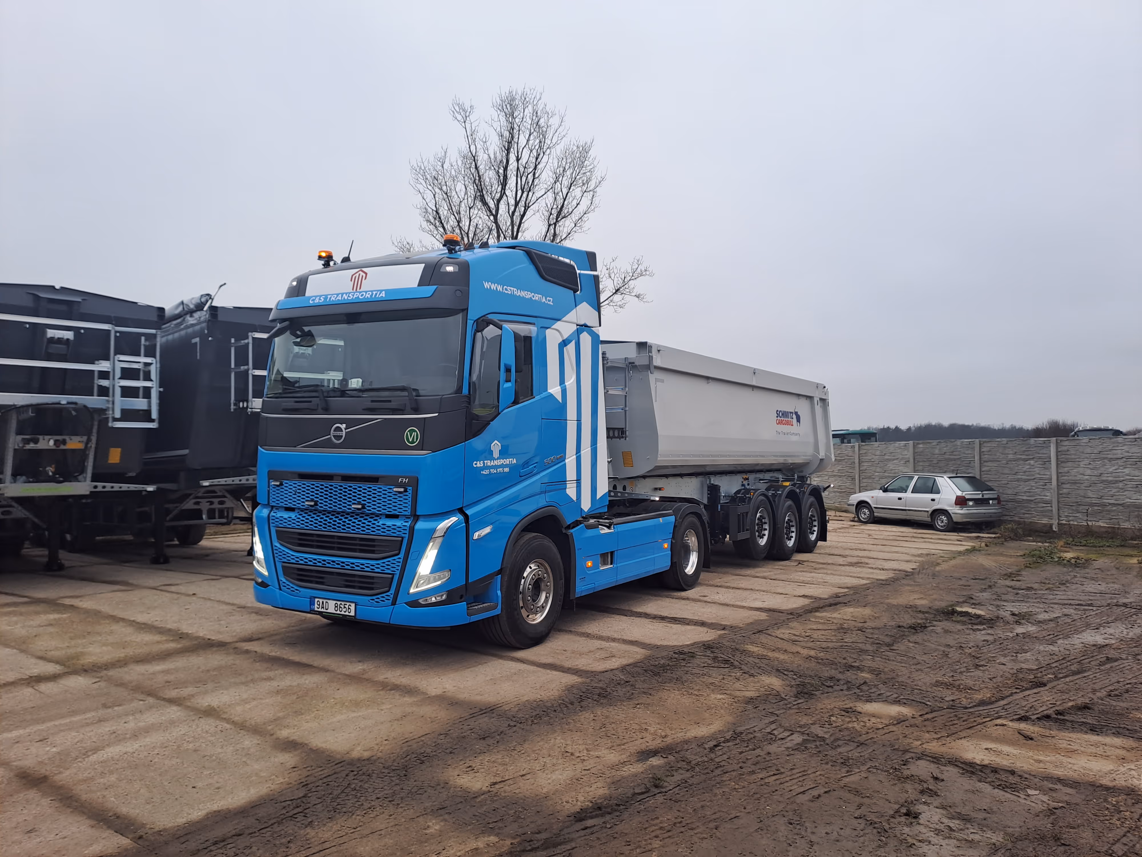 Modrý nákladní tahač Volvo s přívěsem na parkovišti s betonovým povrchem a šedou zdí v pozadí za zatažené oblohy.