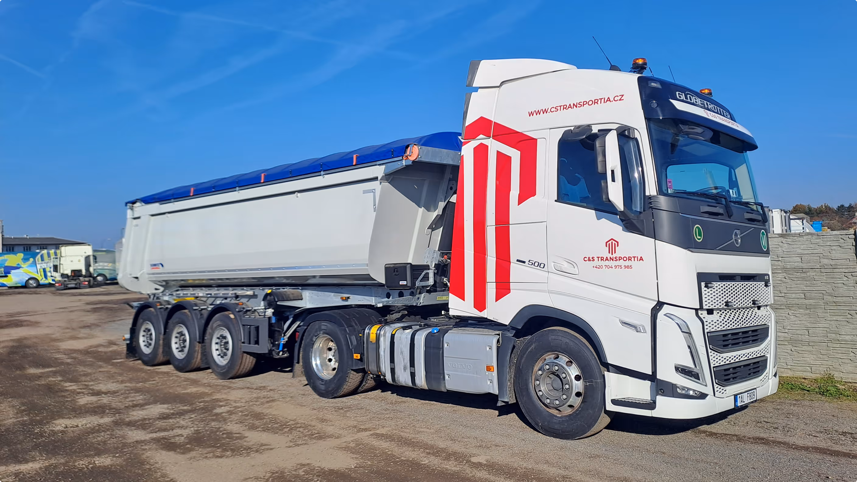 Bílý tahač Volvo s návěsem na silnici za slunečného dne s logem a kontaktními údaji společnosti C&S Transportia.