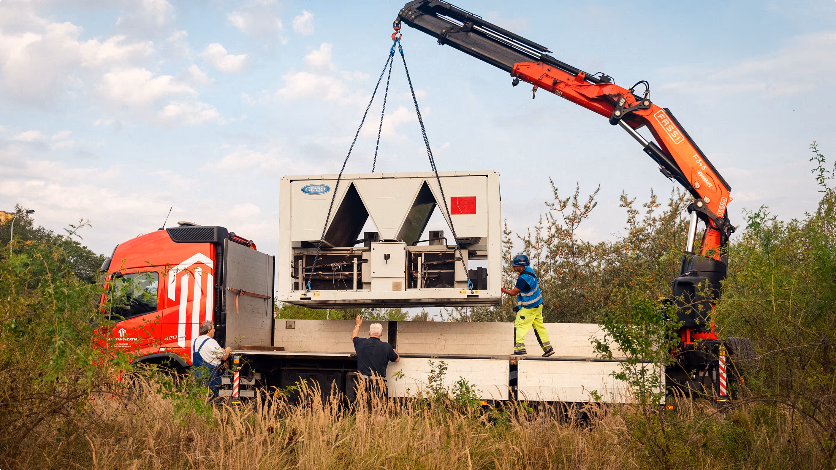 Nákladní červený kamion s jeřábem nakládá velké průmyslové zařízení za pomoci tří dělníků v ochranném oblečení.