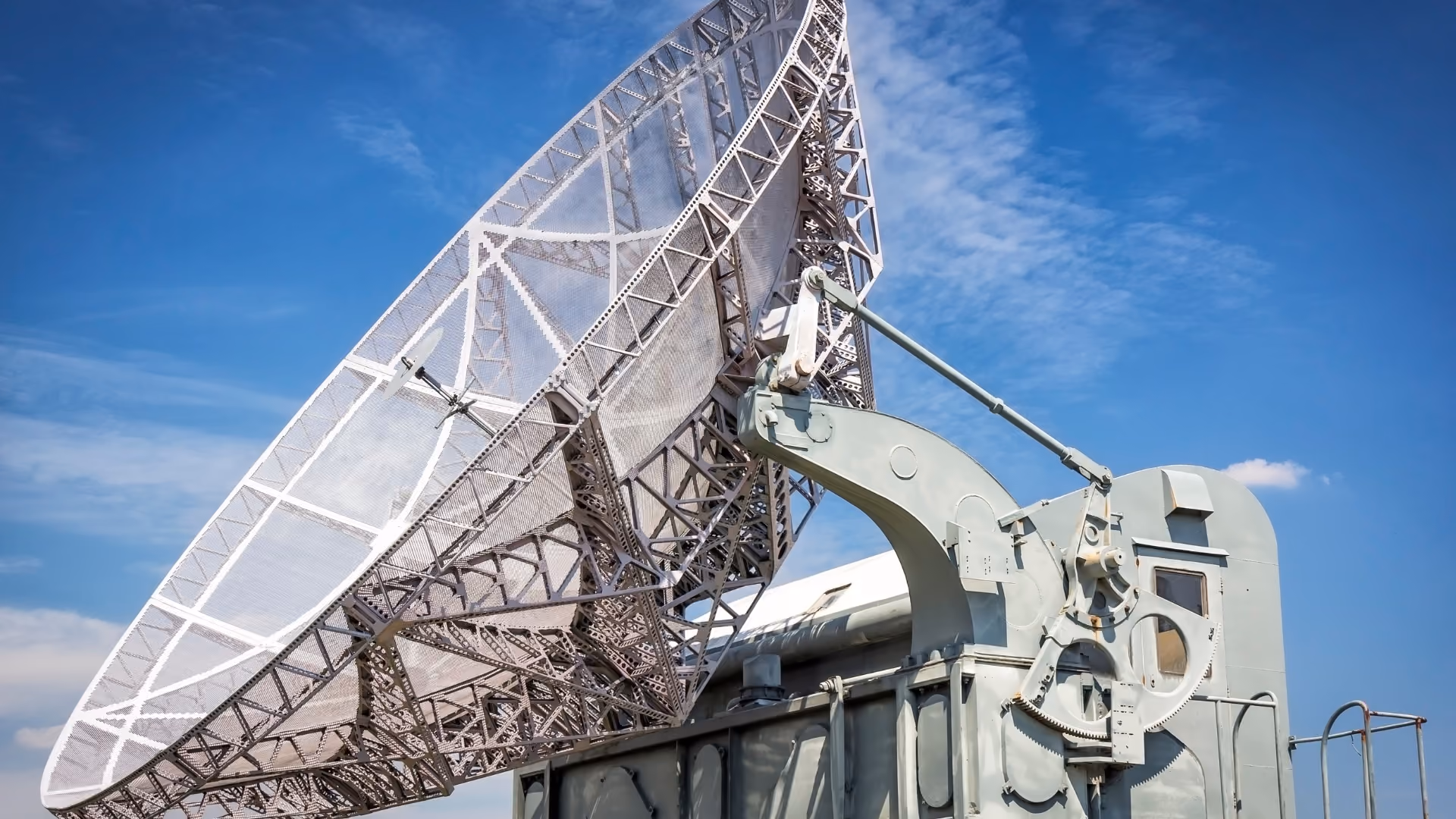 Large metal satellite dish angled upward against a blue sky with scattered clouds.