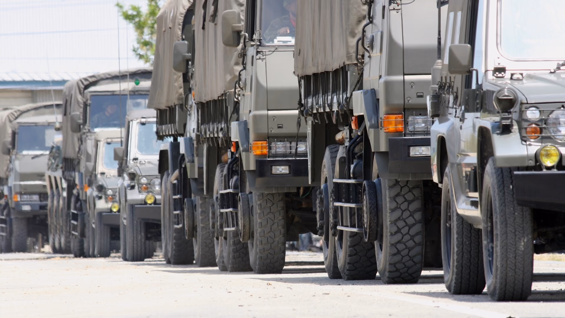 Row of military trucks with canvas covers parked side by side on a dirt road.