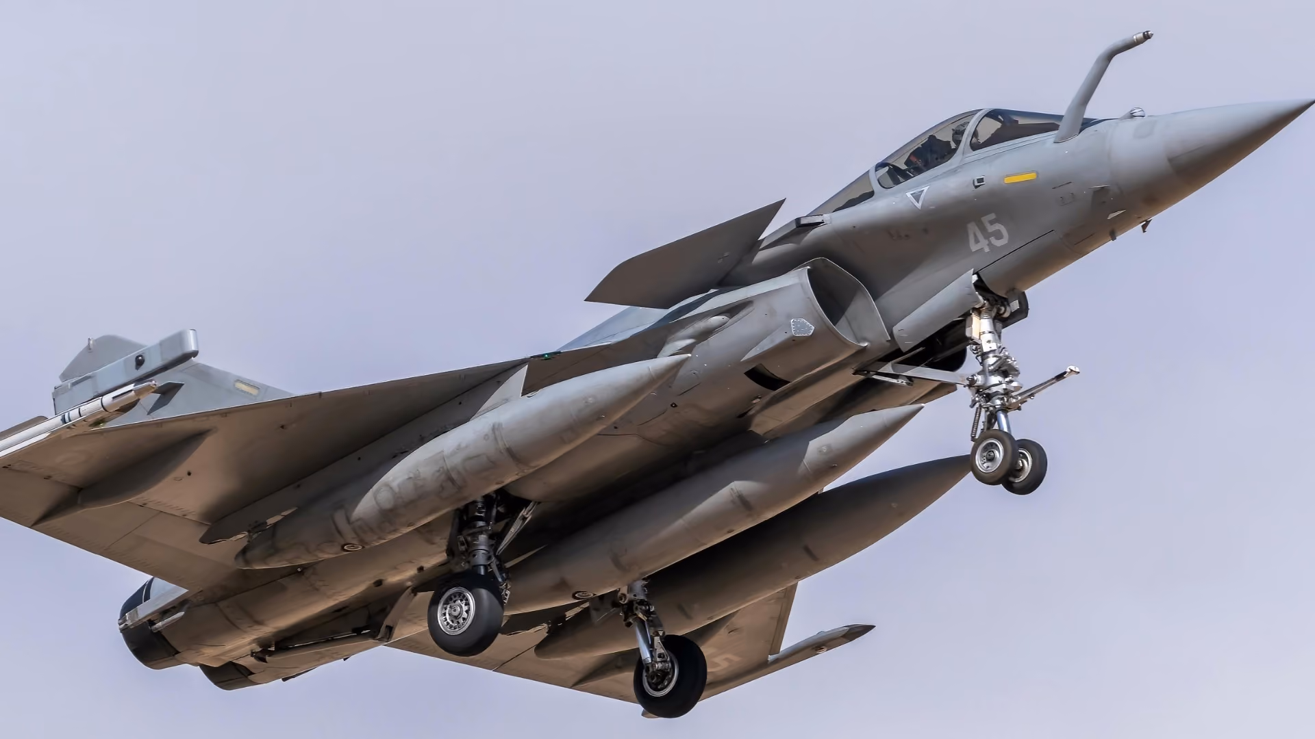 Gray military fighter jet with landing gear extended flying against a clear sky.