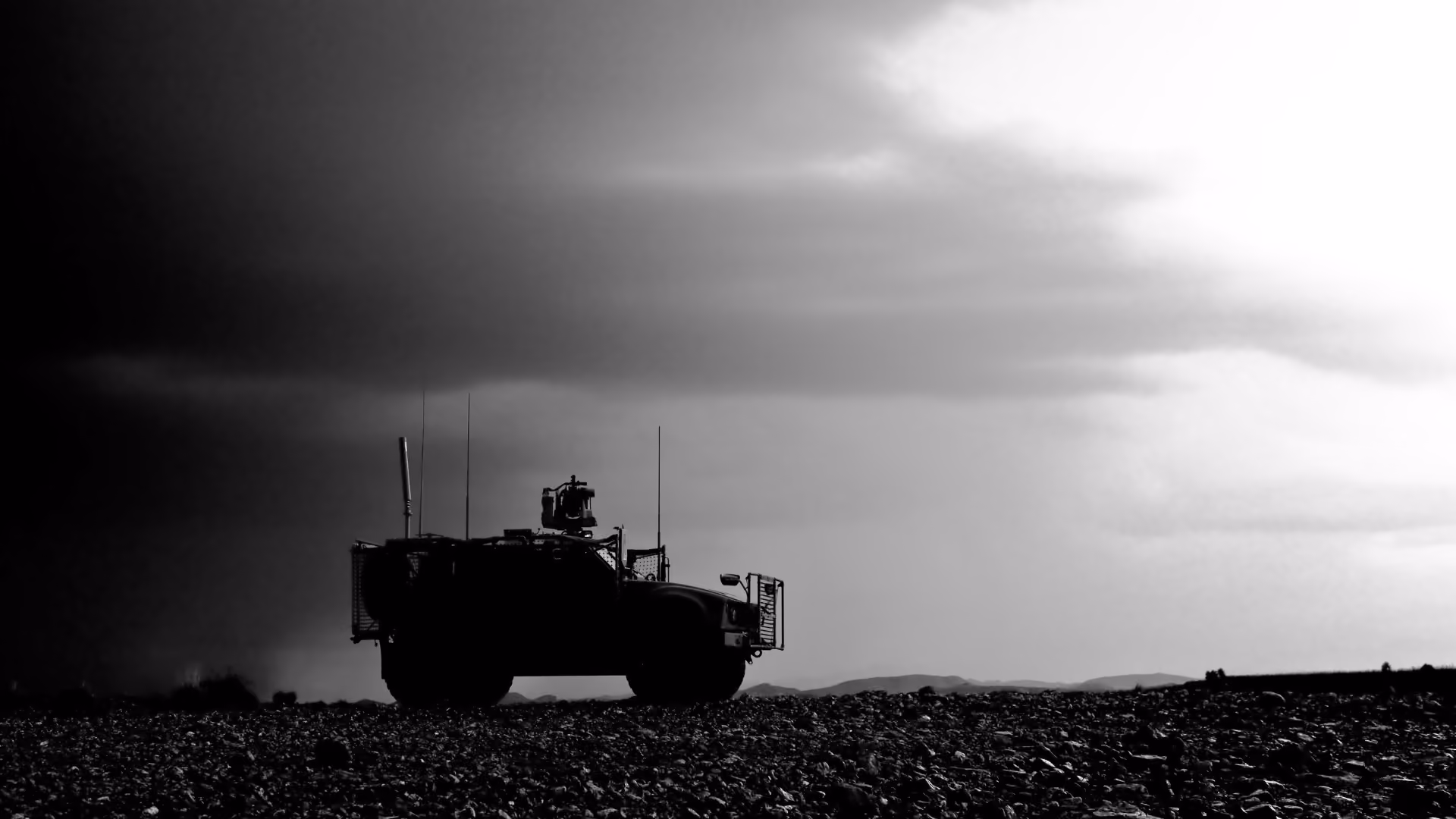 Silhouette of a military vehicle with antennas on rocky terrain under a cloudy sky.