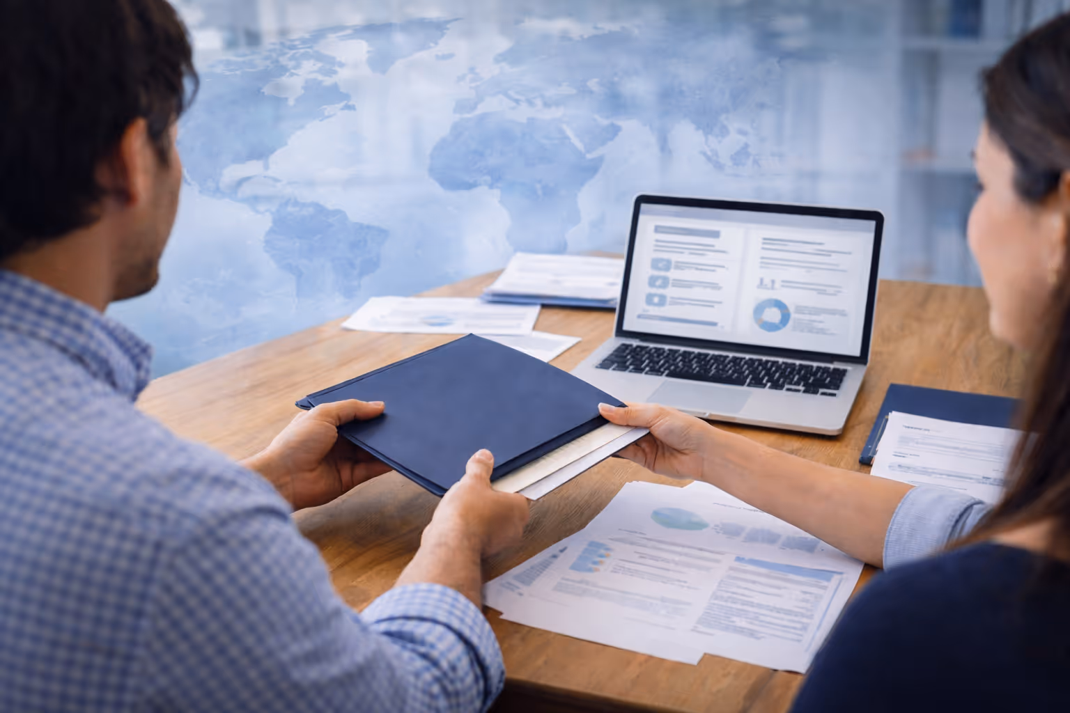 Two business professionals exchange a blue folder over a desk with a laptop and global map background.