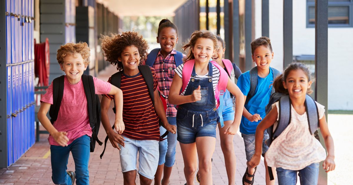 A diverse group of happy elementary school students smiling and running down an outdoor school hallway with backpacks on.