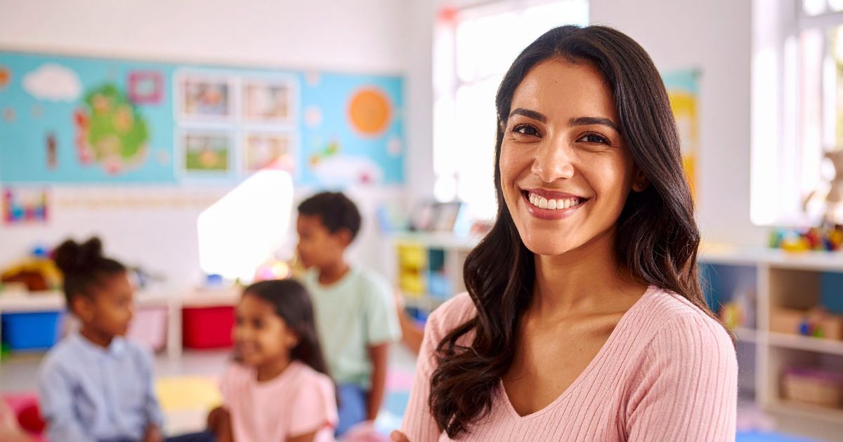 A smiling female international teacher stands in the foreground of a bright American classroom, bringing new perspectives and diverse teaching methodologies to U.S. students.