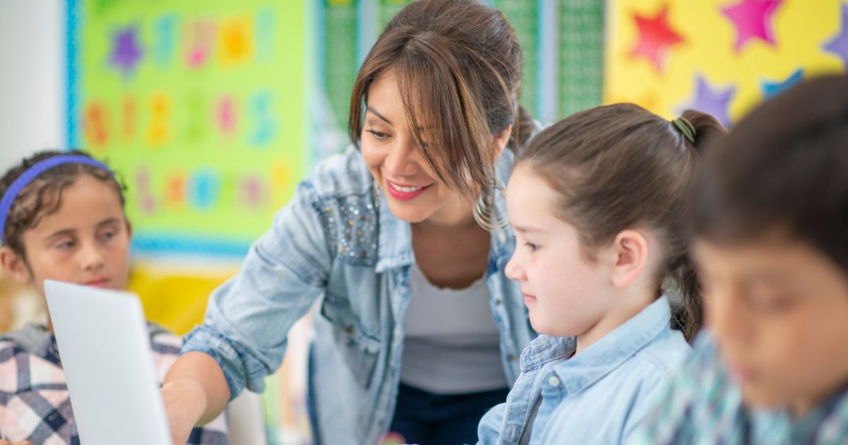 An international teacher smiles as she guides an elementary school student on a laptop in a bright classroom, demonstrating a global perspective in education.