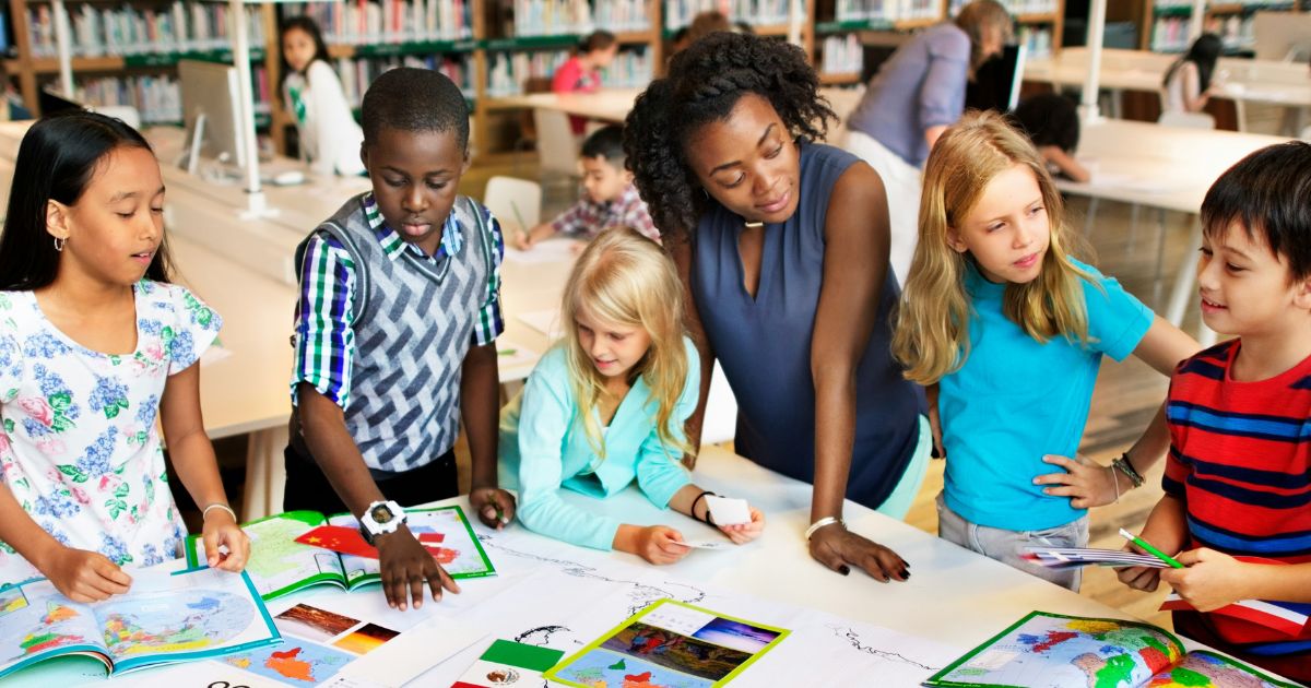 An international teacher guides a diverse group of elementary school students working together at a table covered in maps and cultural materials in a library.
