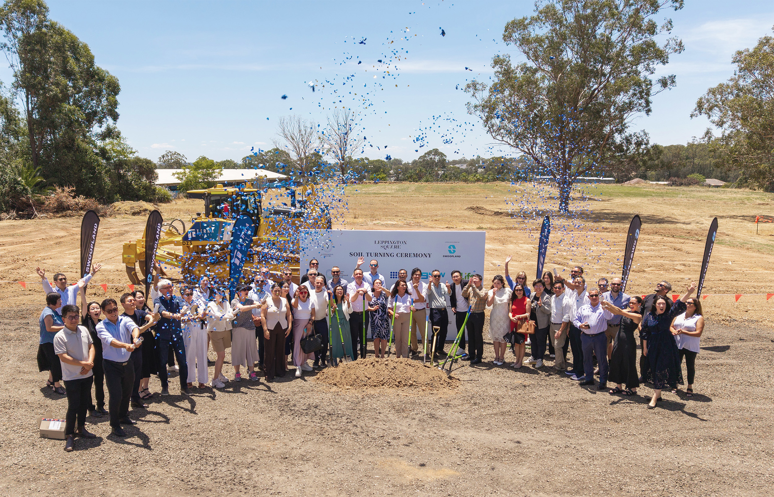 Group photo of Swoopland team and stakeholders at the Rickard Gardens soil turning ceremony, wearing high-visibility vests and holding shovels, with Swoopland banners in the background