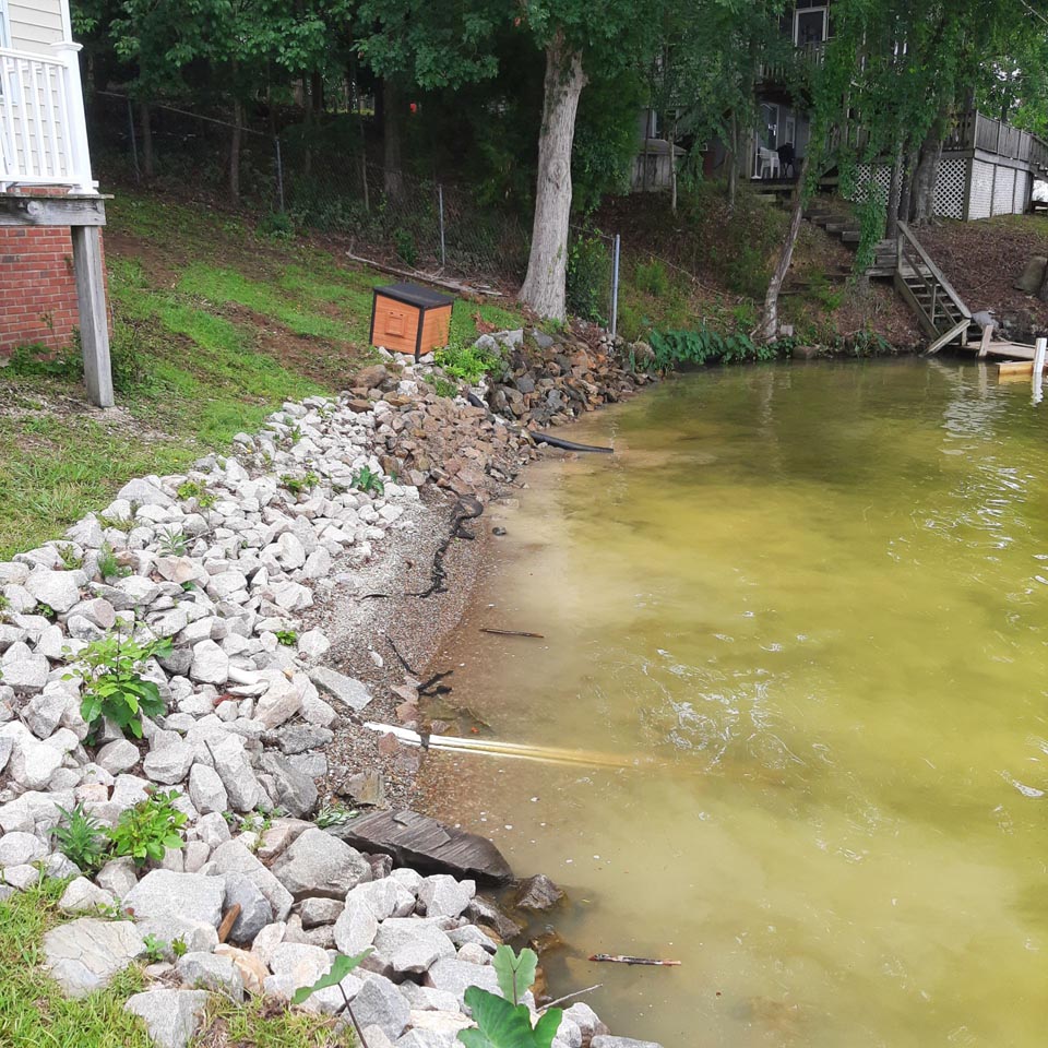 A dirty shoreline with different colored rocks, sand, and weeds. The shore has receded from being unmanaged.