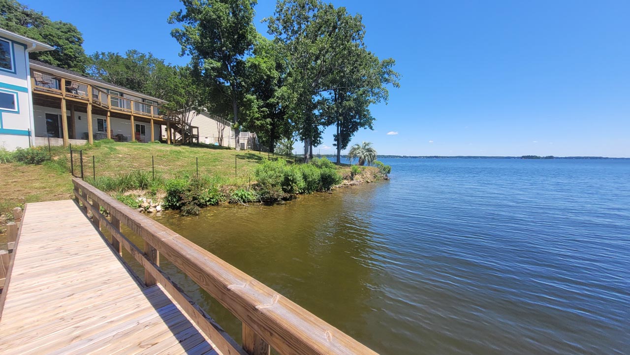 A view of the lakeshore from a dock, unmanaged shoreline with bushes and weeds.