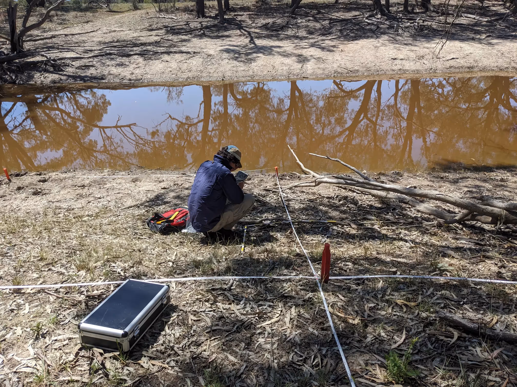 Person crouching down by water's edge with back to camera and white tape on ground making a cross. 