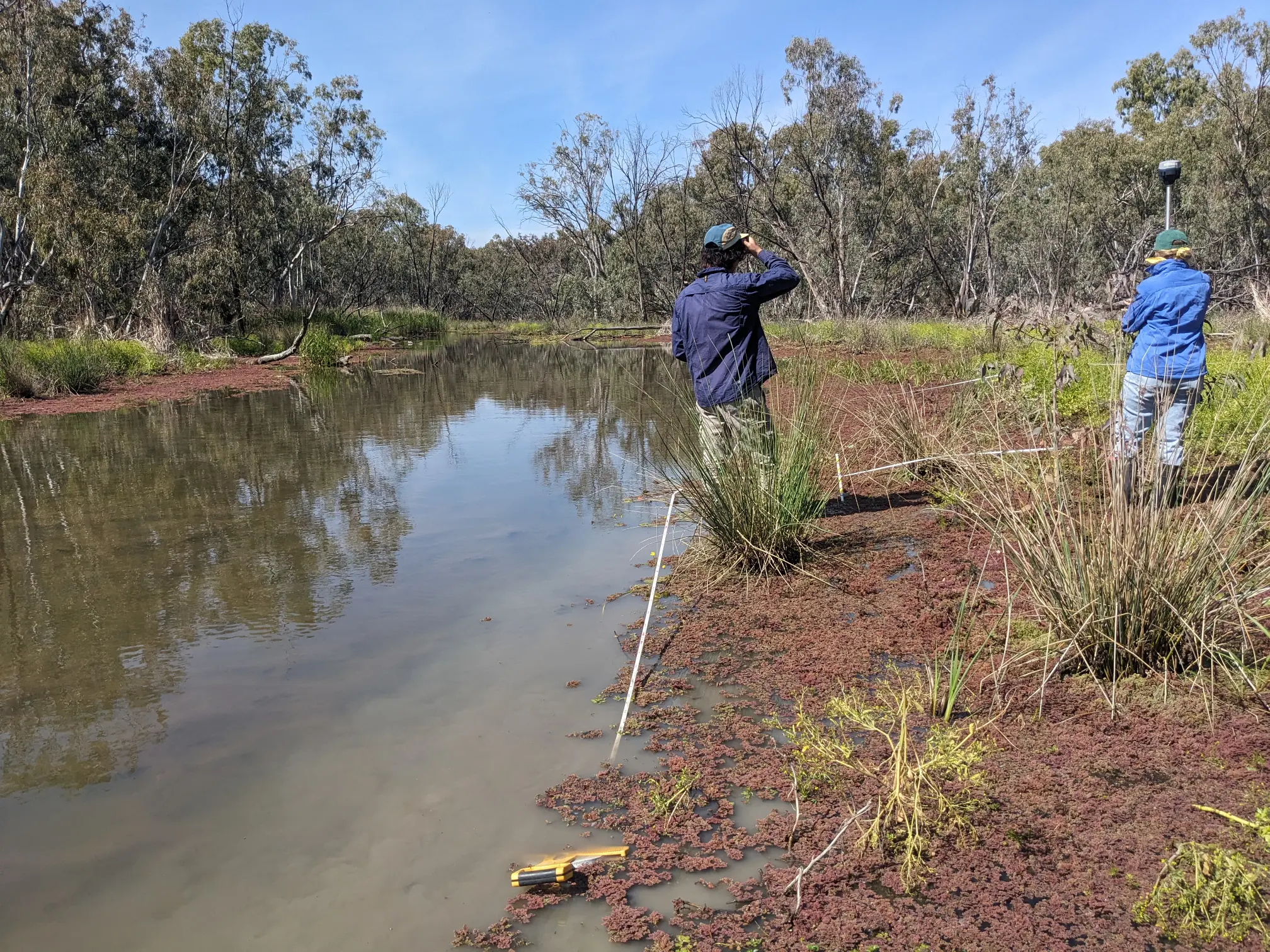 Two people standing next to a waterway. One person holding a tall pole.