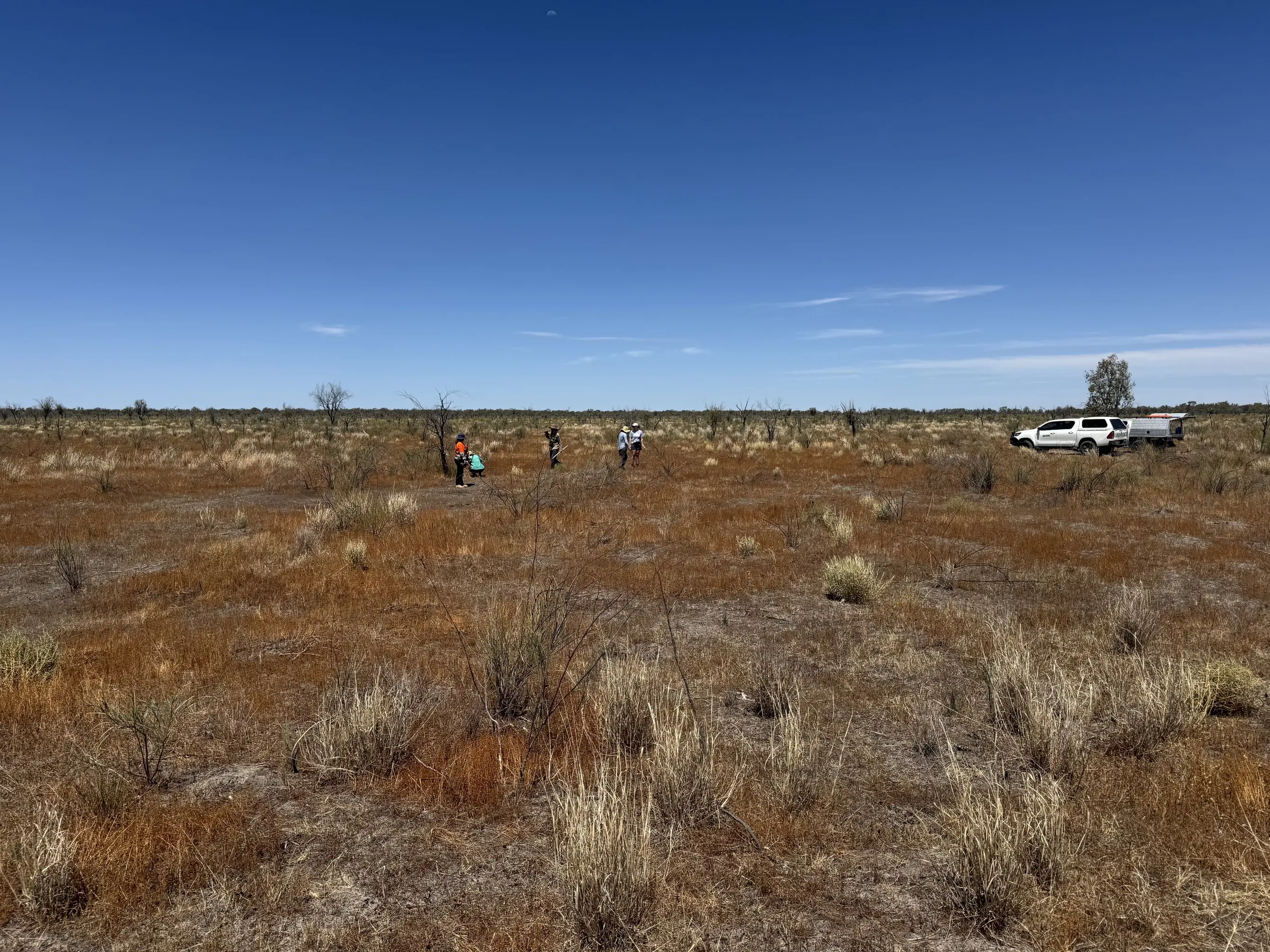 Dry shrubland with vast sky behind. Several people in the distance and a vehicle.