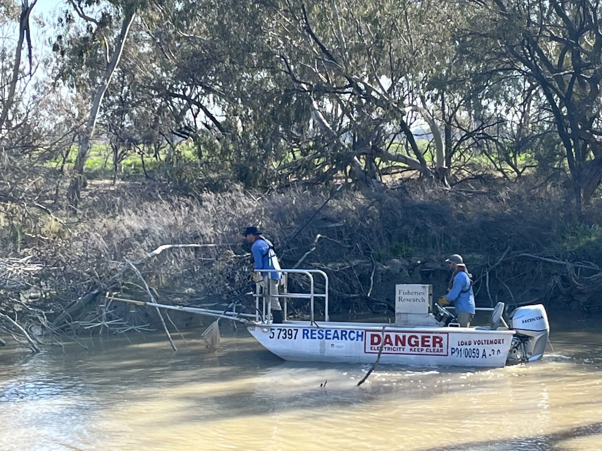 Picture of two people in a medium sized fishing boat on the Macquarie River with brown water and riverbank in the background. One person is at the bow of the boat fishing and the other is driving the boat.