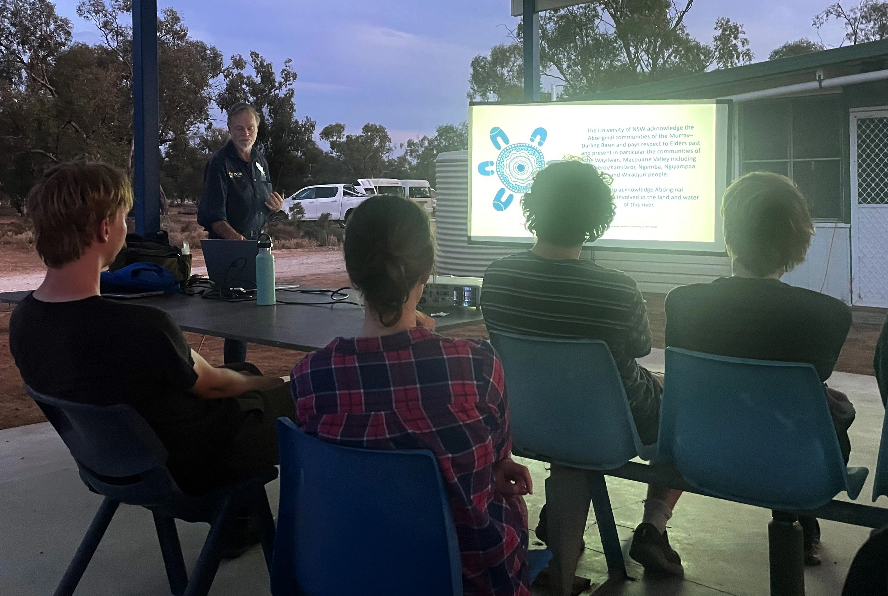 A person standing beside a screen presenting and the backs of the audience's heads 