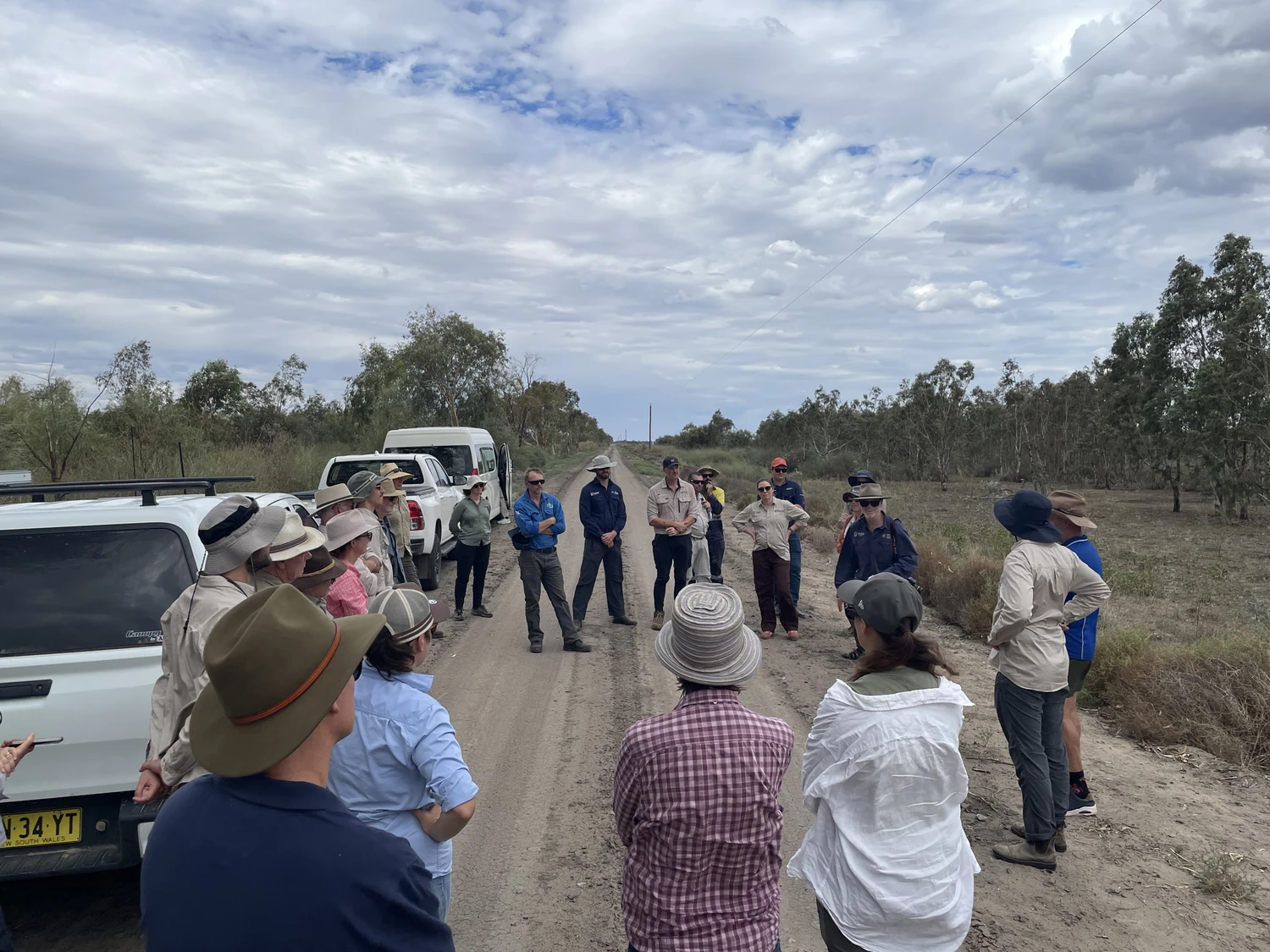 A group of people standing on a dirt road in a rural area, engaged in a discussion. Several vehicles are parked nearby.