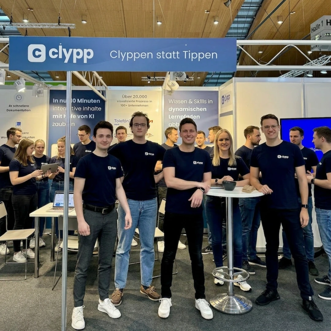 Group of people wearing matching navy blue clypp t-shirts posing at a trade show booth with a blue sign overhead reading 'clypp Clyppen statt Tippen'.
