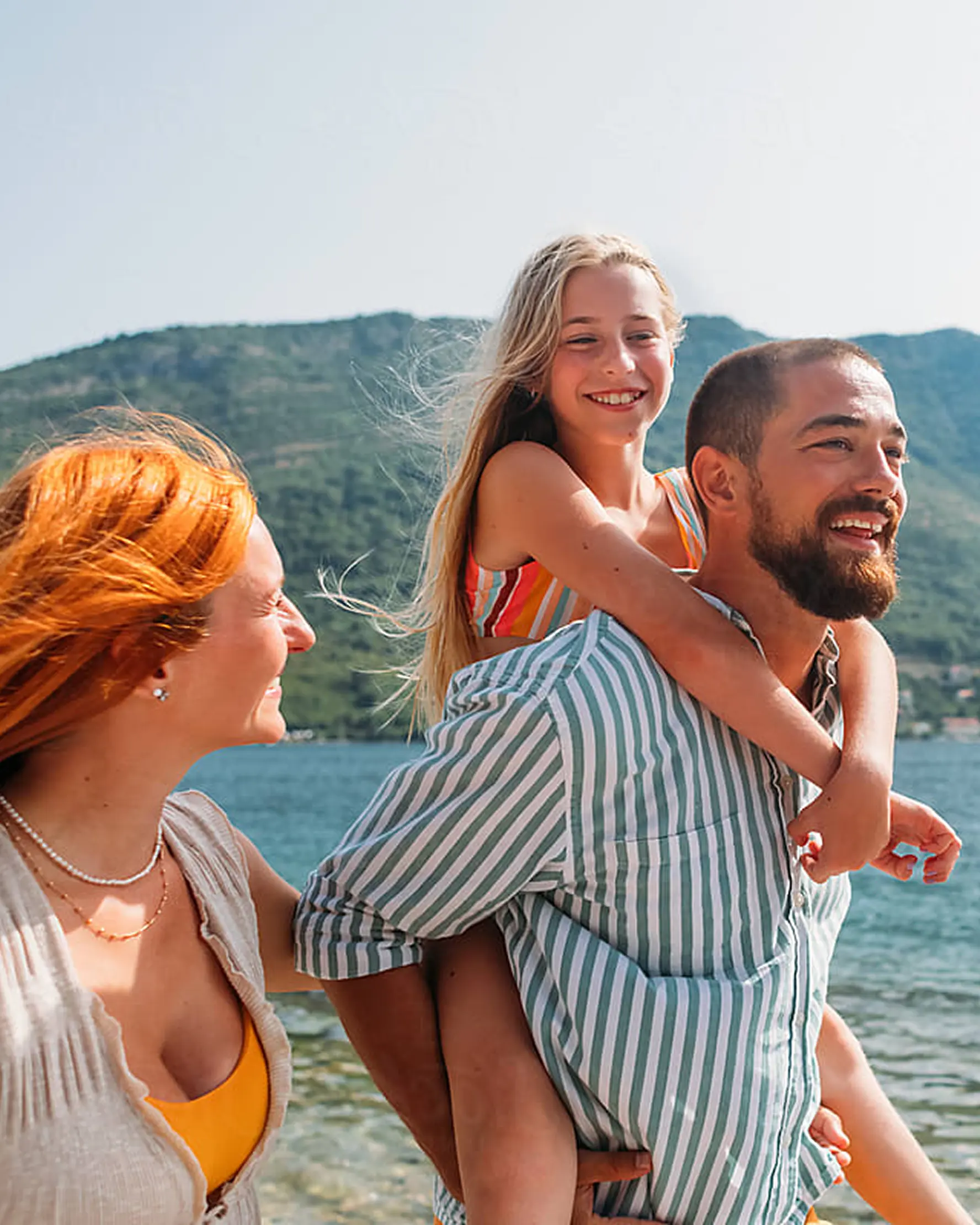 A joyful family by the lake, with a man carrying a smiling girl on his back and a woman with flowing red hair. Mountains and water in the background.