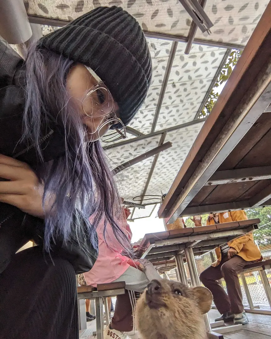 Woman with glasses and purple hair sits under a patterned canopy, facing a small quokka looking up at her. People are seated at tables in the background.
