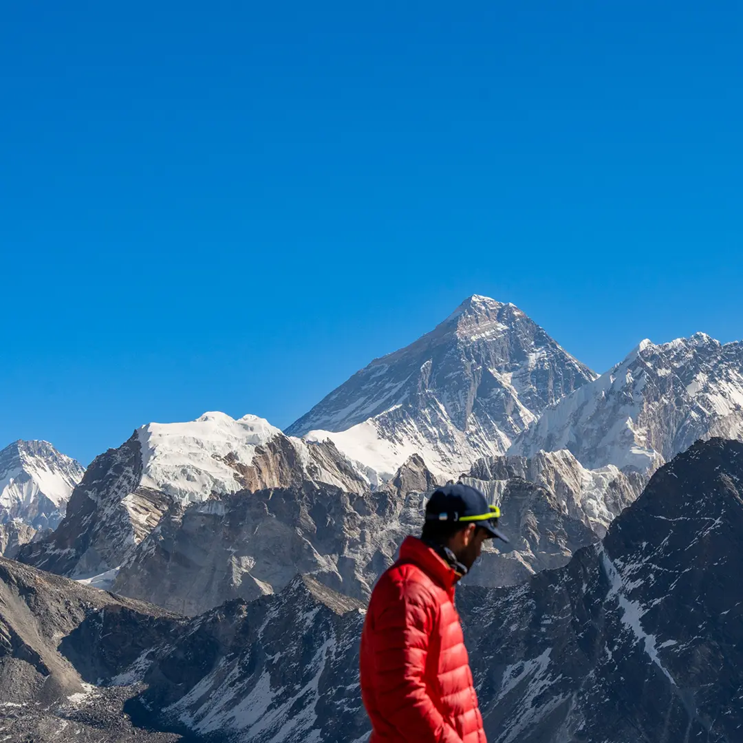 A person in a red jacket stands against a backdrop of snow-capped mountains with a clear blue sky, evoking a sense of adventure and tranquility.