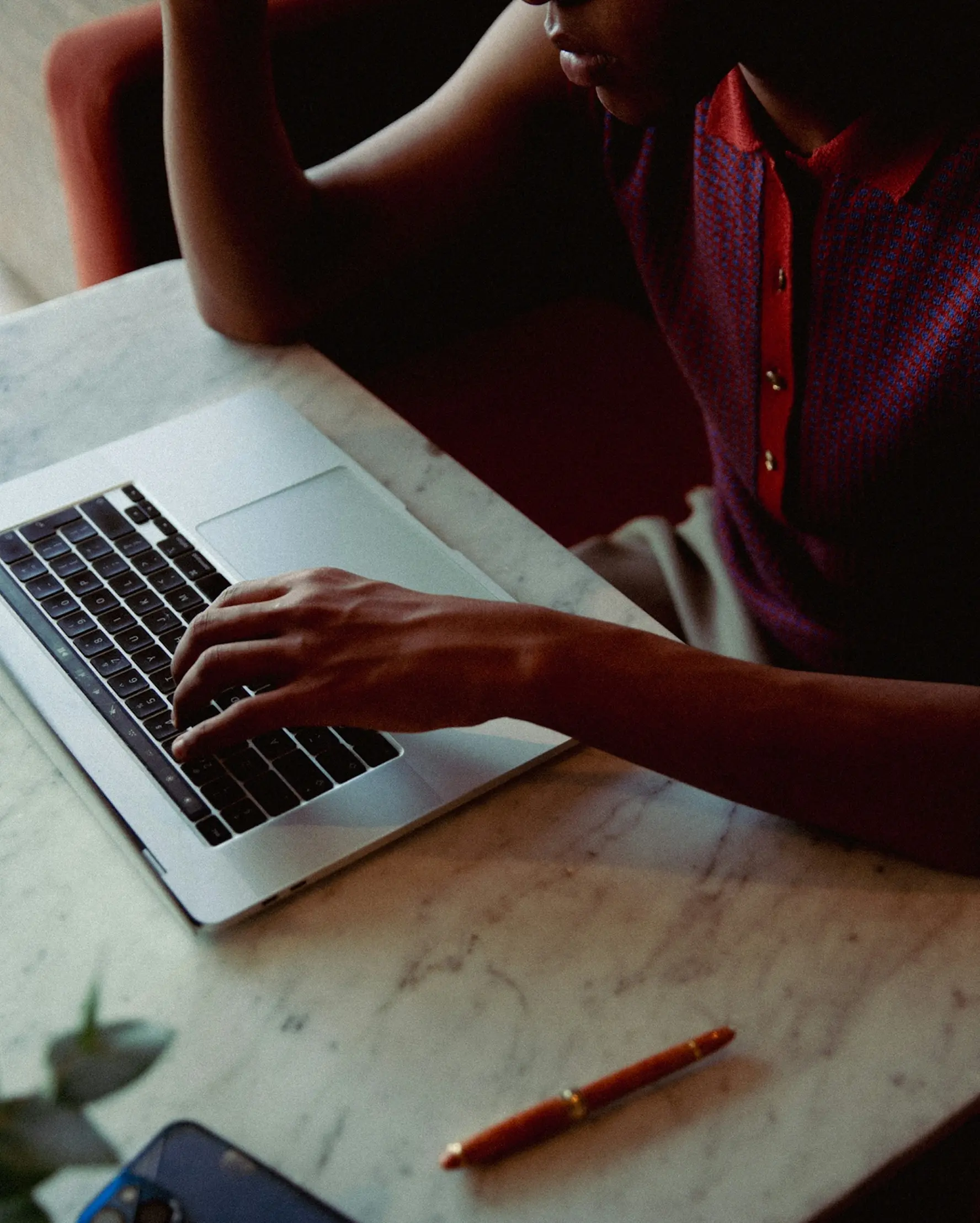 Person in a red and pink shirt, types on a laptop at a marble table. A pen and phone are nearby. The scene conveys focus and productivity.