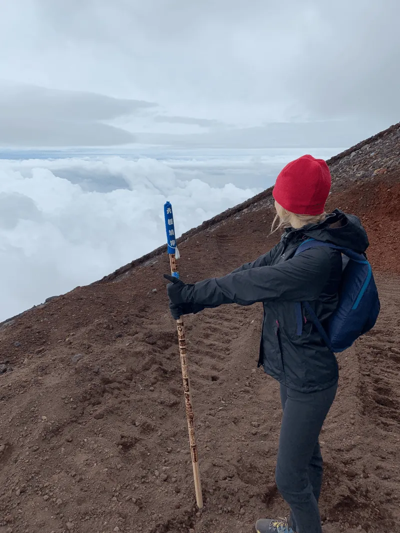 A person in a red beanie and black jacket stands on a rocky mountain slope, holding a wooden stick. They're overlooking a vast sea of clouds under a gray sky, conveying a sense of adventure and tranquility.