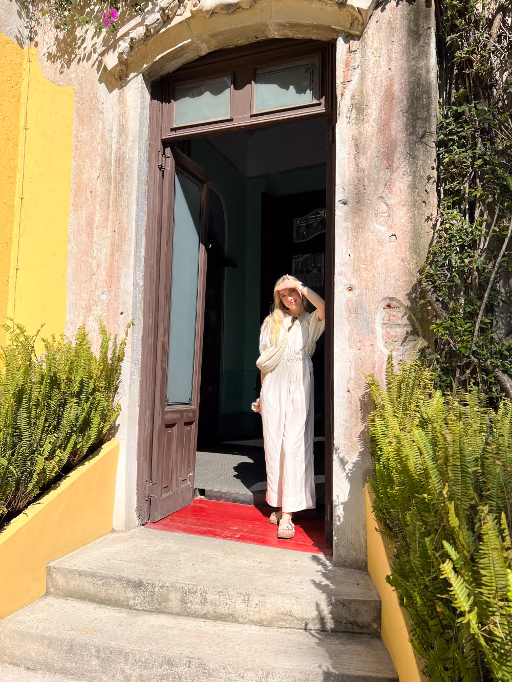 Woman in a white dress stands in a doorway, shielding her eyes from the sun. Bright yellow walls and green ferns create a vibrant, cheerful atmosphere.