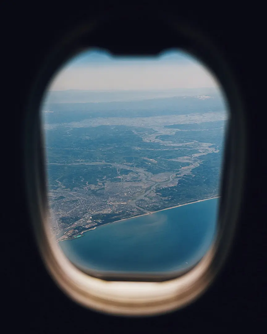 Aerial view from an airplane window showing a coastal city with winding rivers and sprawling landscape. The scene conveys a calm, expansive horizon.